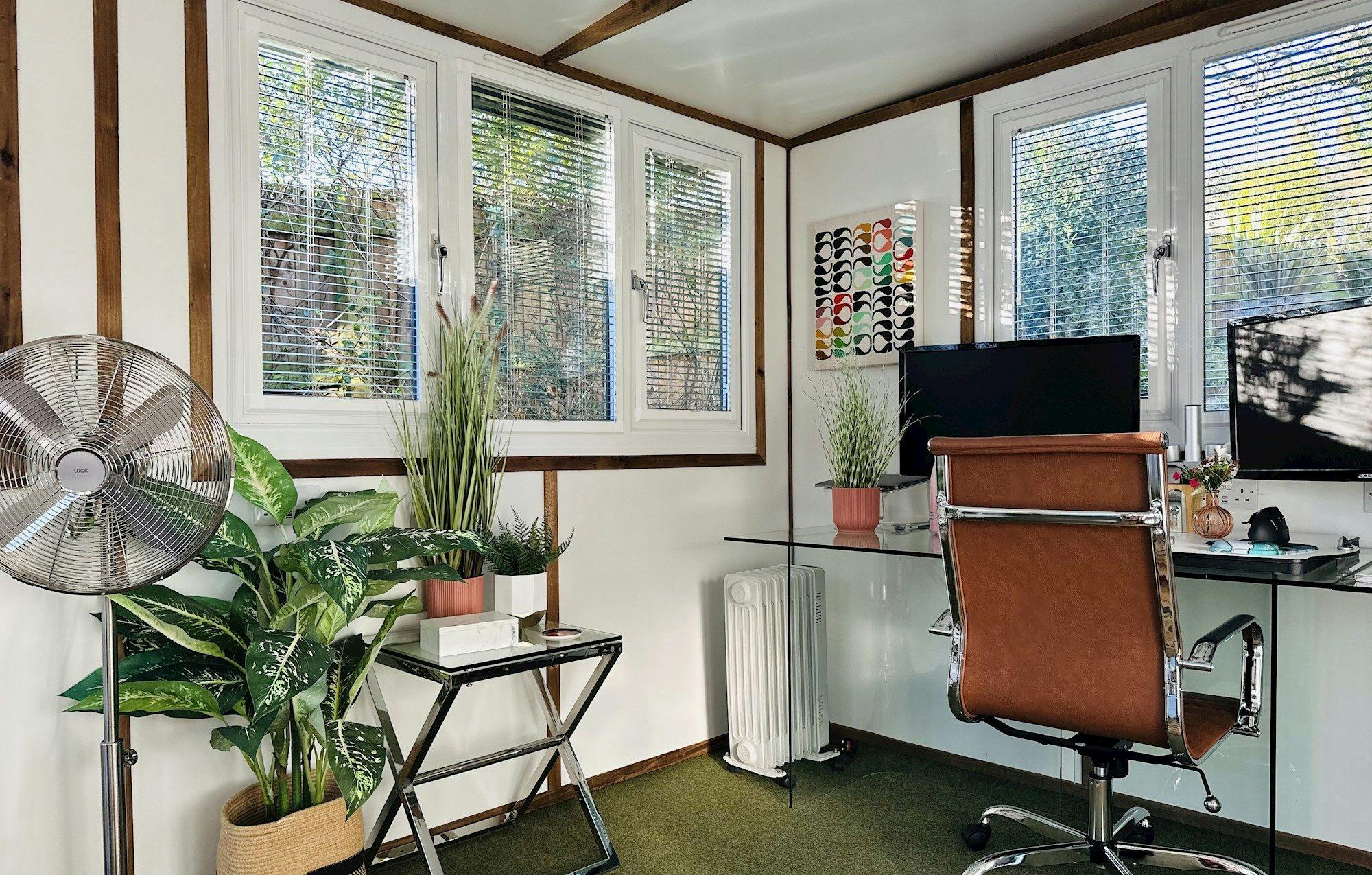 A modern home office features a brown chair facing dual monitors on a glass desk. Potted plants and an electric fan enhance the airy space, surrounded by large windows and green carpet.