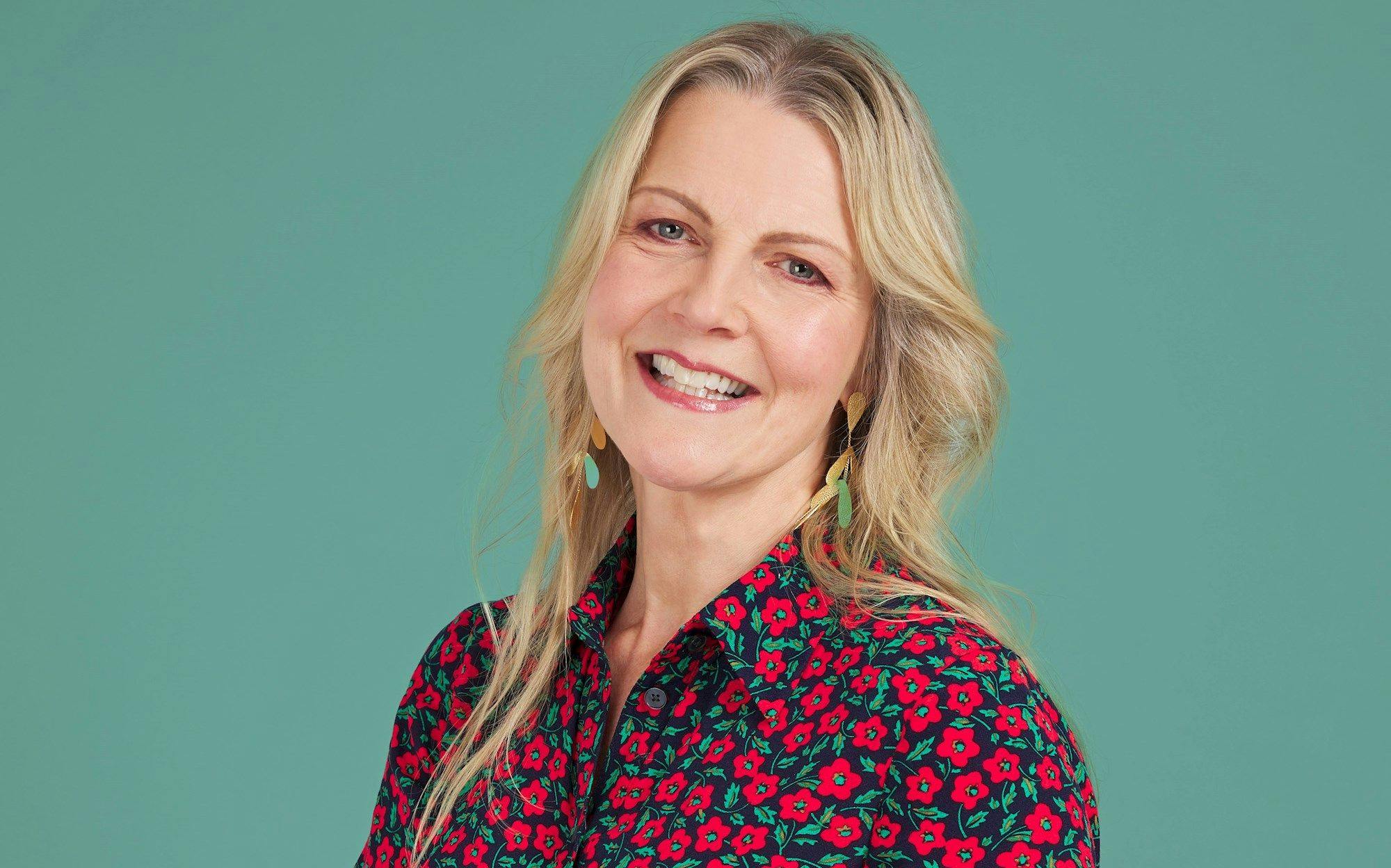 A person smiles, wearing a colorful floral shirt and long earrings, against a teal background.