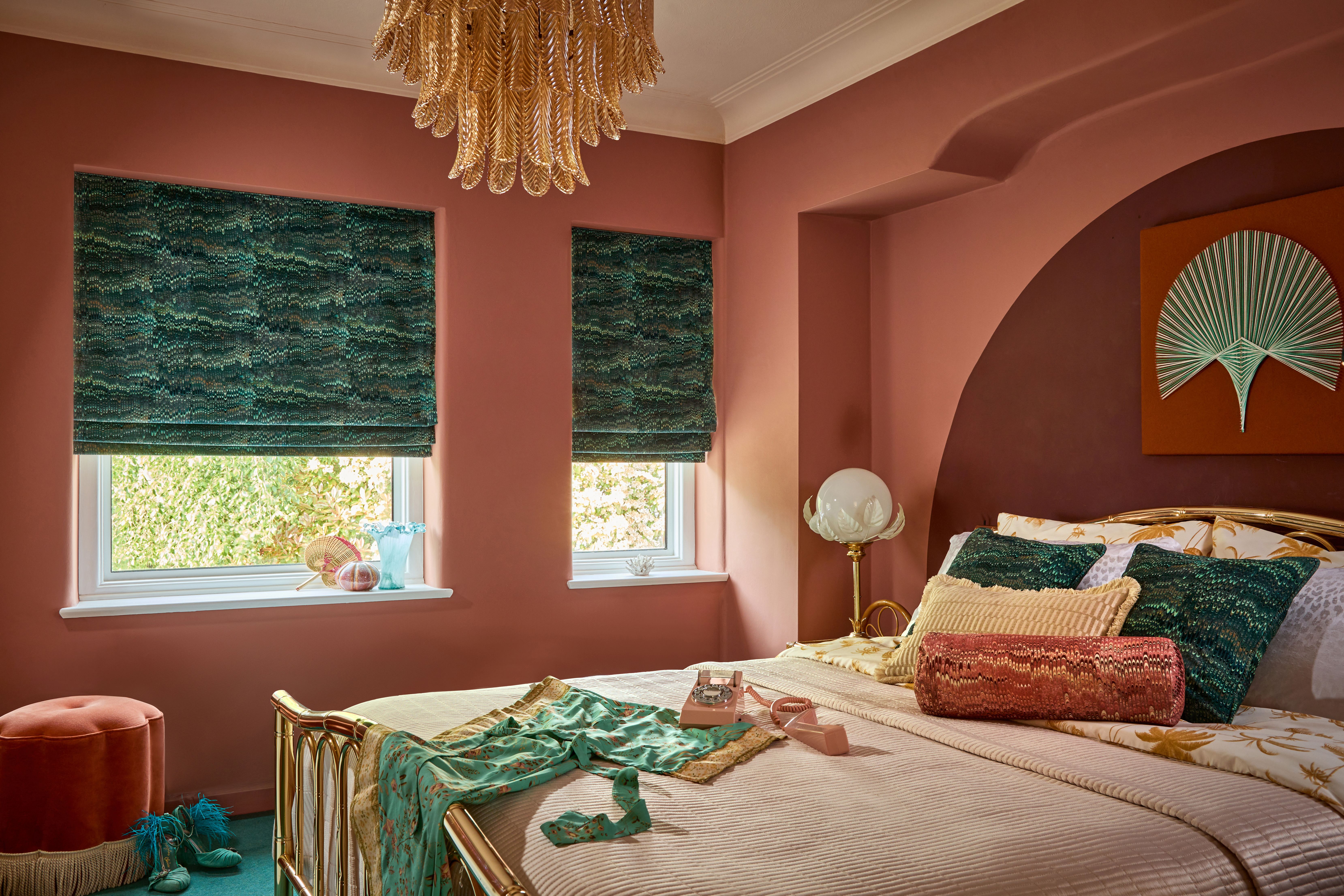 A bedroom features a bed with patterned pillows and a vintage phone, situated against pink walls with green blinds. A decorative chandelier hangs above, adding elegance to the setting.