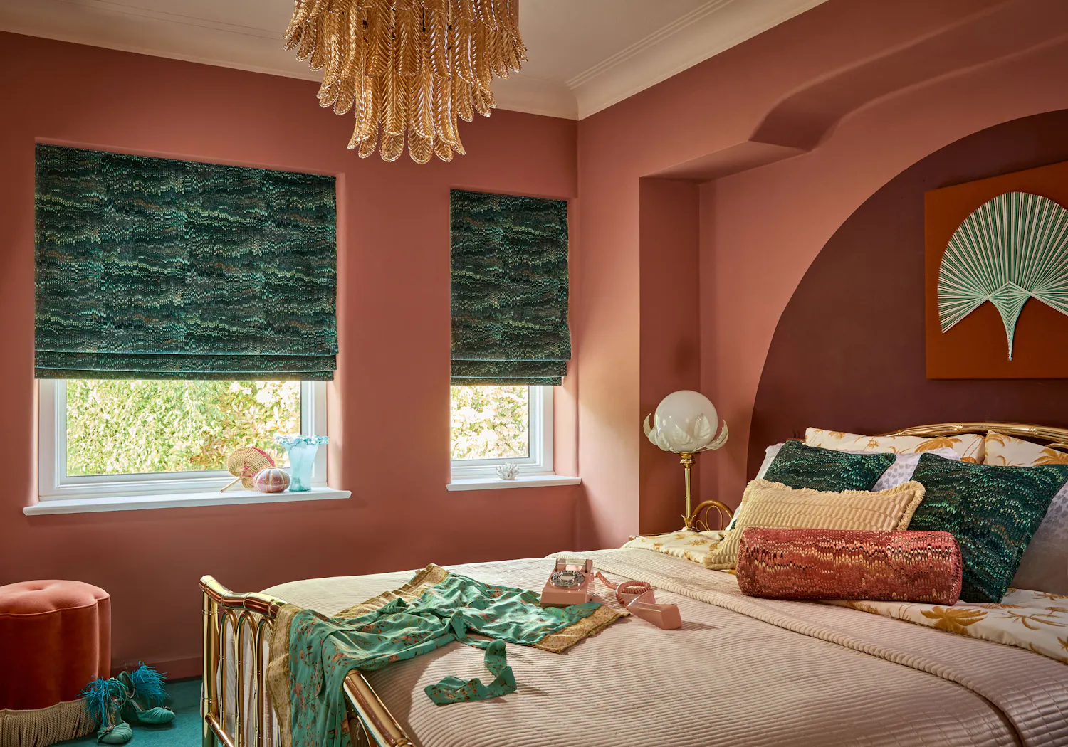 A bedroom features a bed with patterned pillows and a vintage phone, situated against pink walls with green blinds. A decorative chandelier hangs above, adding elegance to the setting.
