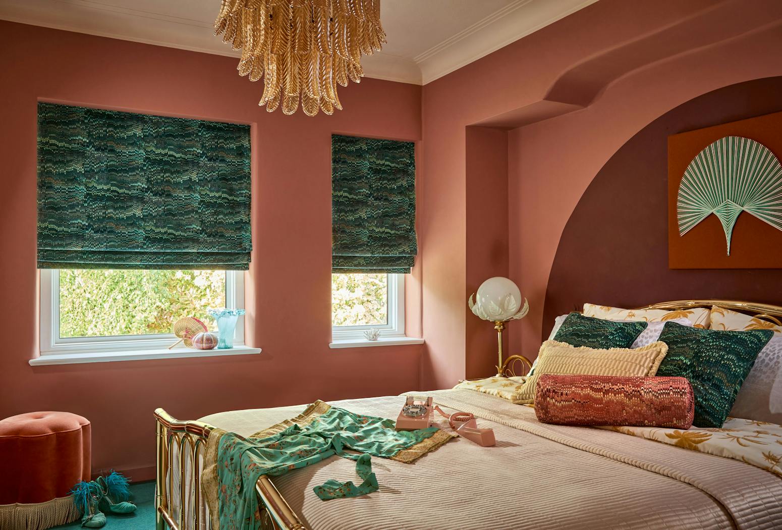 A bedroom features a bed with patterned pillows and a vintage phone, situated against pink walls with green blinds. A decorative chandelier hangs above, adding elegance to the setting.