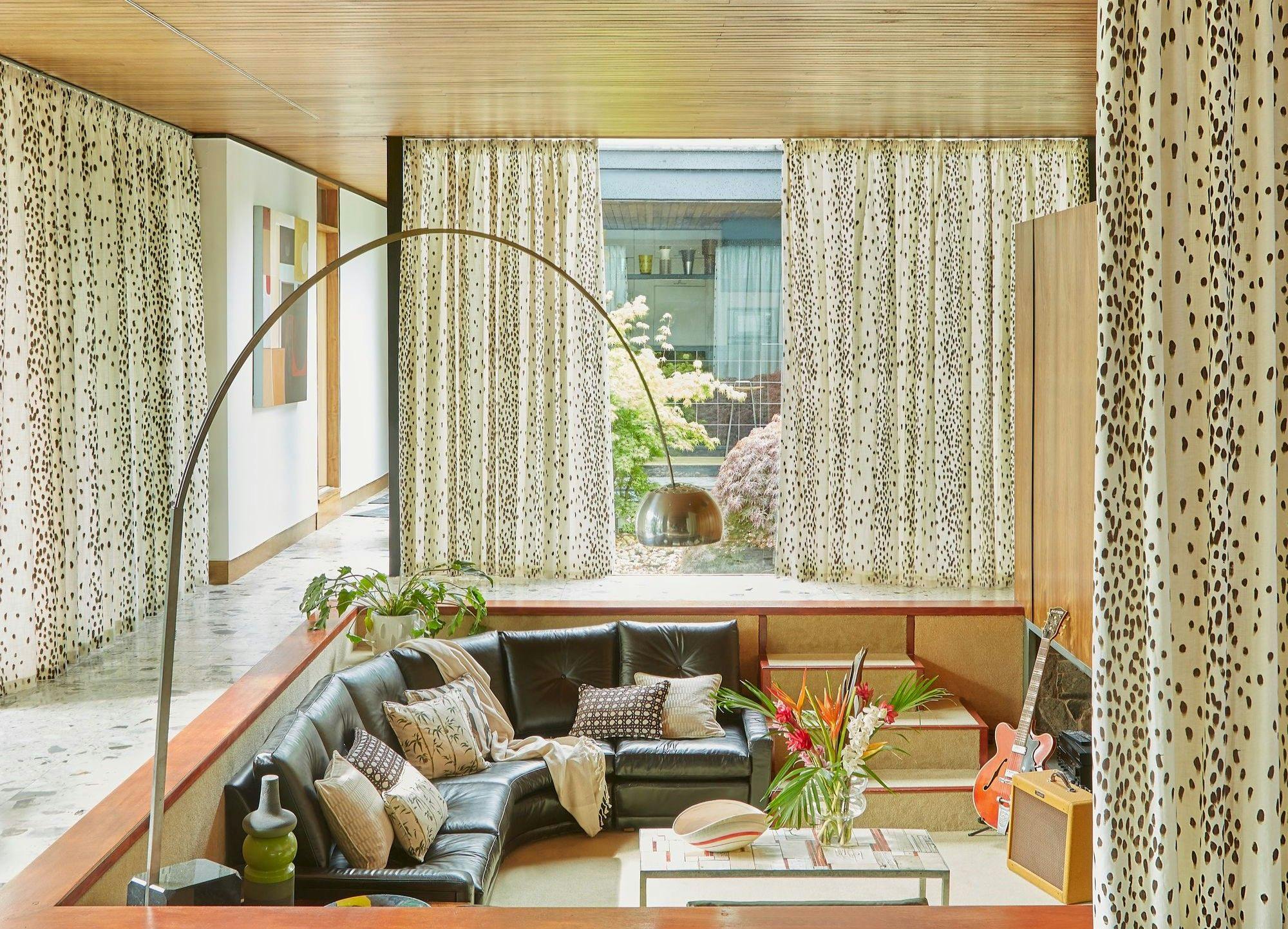 A black leather sectional couch, adorned with various patterned pillows, is set in a cozy living room. Surrounding it are spotted curtains, a modern floor lamp, and indoor plants.