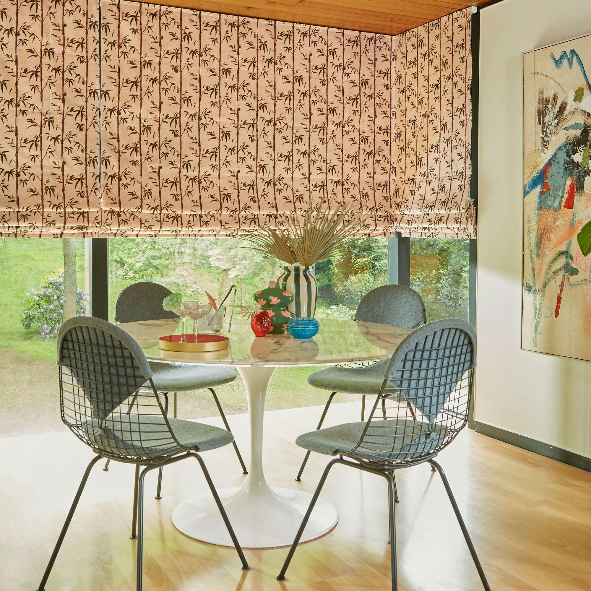 A round table with decorative vases and glassware is surrounded by wire chairs in a room with patterned curtains and abstract art. Large windows overlook a lush garden.