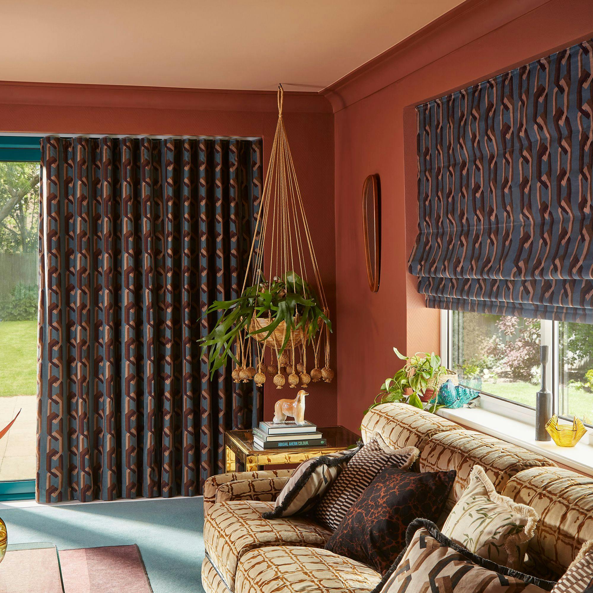 A patterned sofa with assorted cushions sits in a living room. Curtains match the sofa's geometric design. A hanging plant decorates one corner, with sunlight streaming through a window.