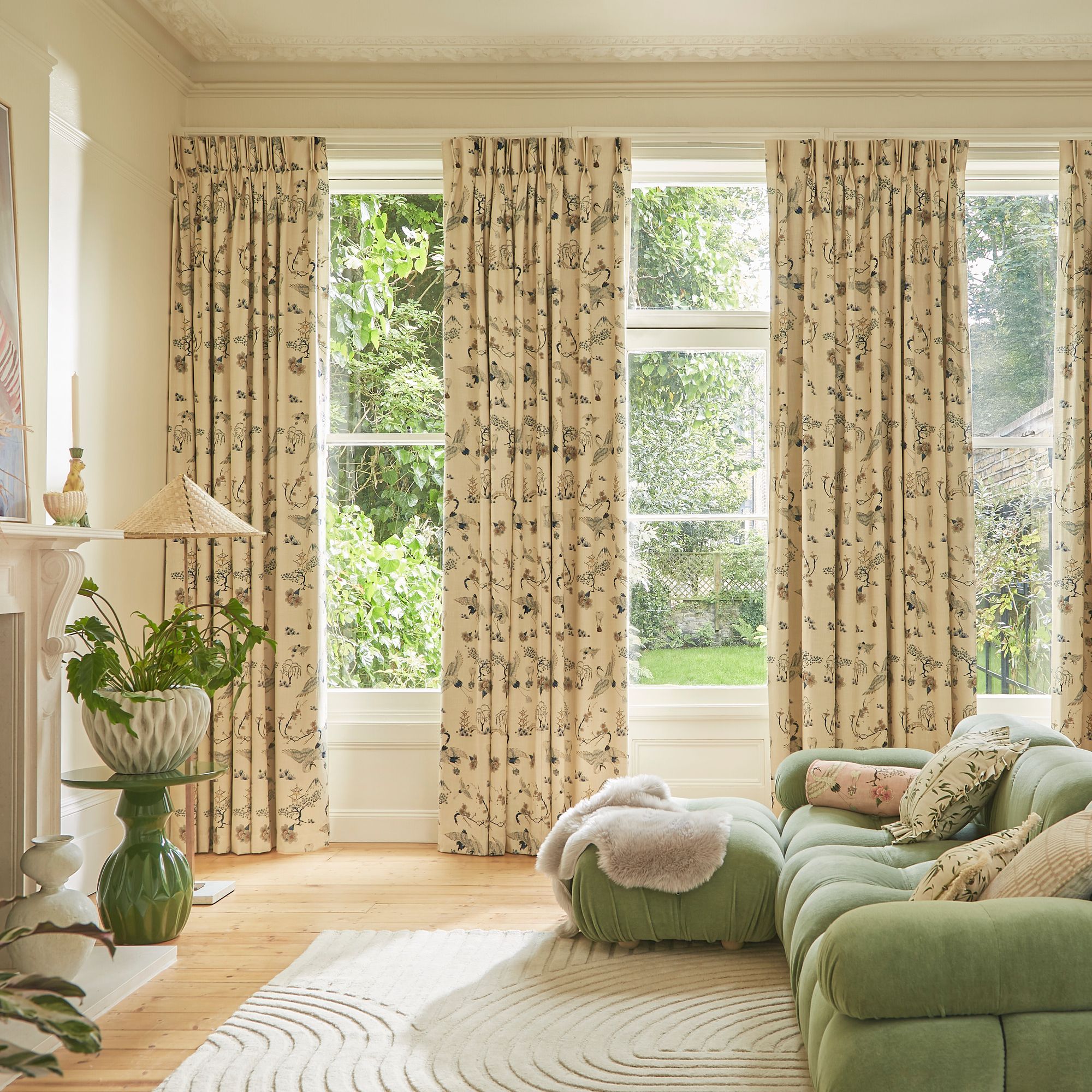 A green sofa with cushions sits in a bright living room with floral curtains. Sunlight streams through large windows, illuminating a potted plant on a side table and a white rug.
