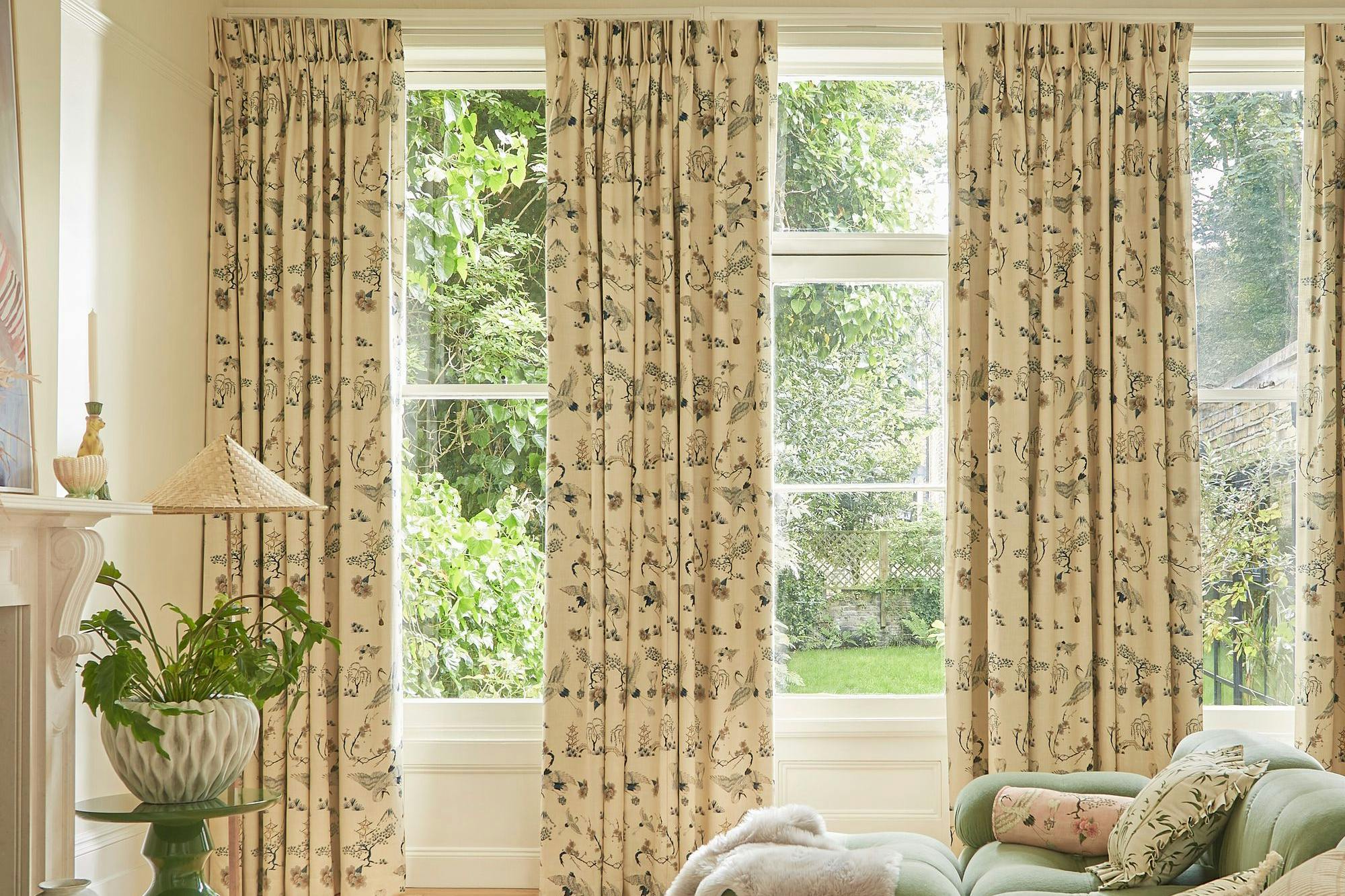 A green sofa with cushions sits in a bright living room with floral curtains. Sunlight streams through large windows, illuminating a potted plant on a side table and a white rug.