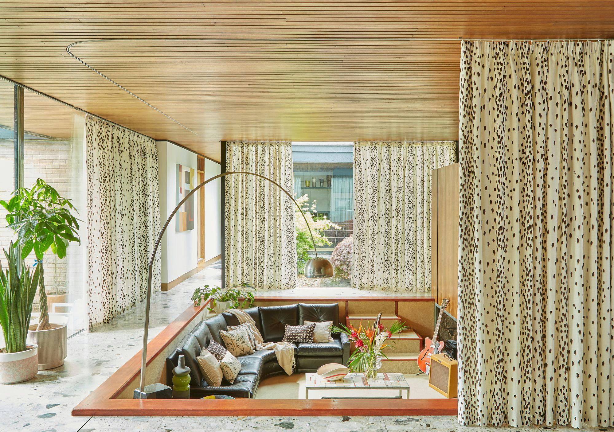 Sunken black leather sofa with patterned cushions sits centrally, surrounded by dotted curtains. A large plant and decorative floor lamp accent the modern, spacious room with wooden ceiling.