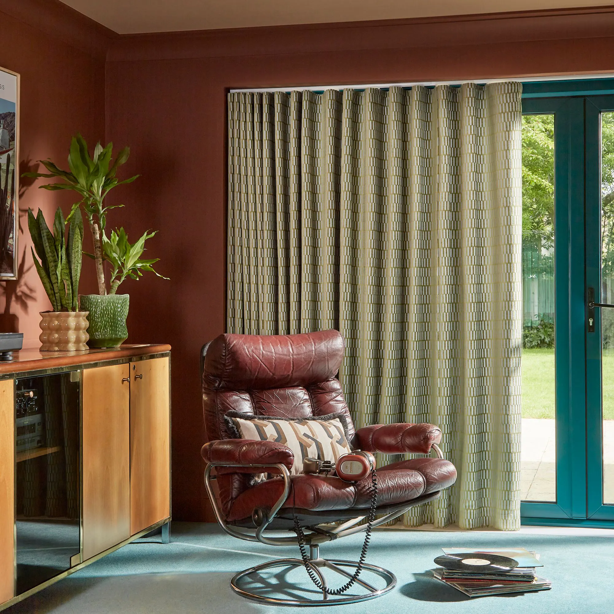 A brown leather armchair sits beside a stack of vinyl records on a carpeted floor, with potted plants on a wooden cabinet and sunlight streaming through patterned curtains.