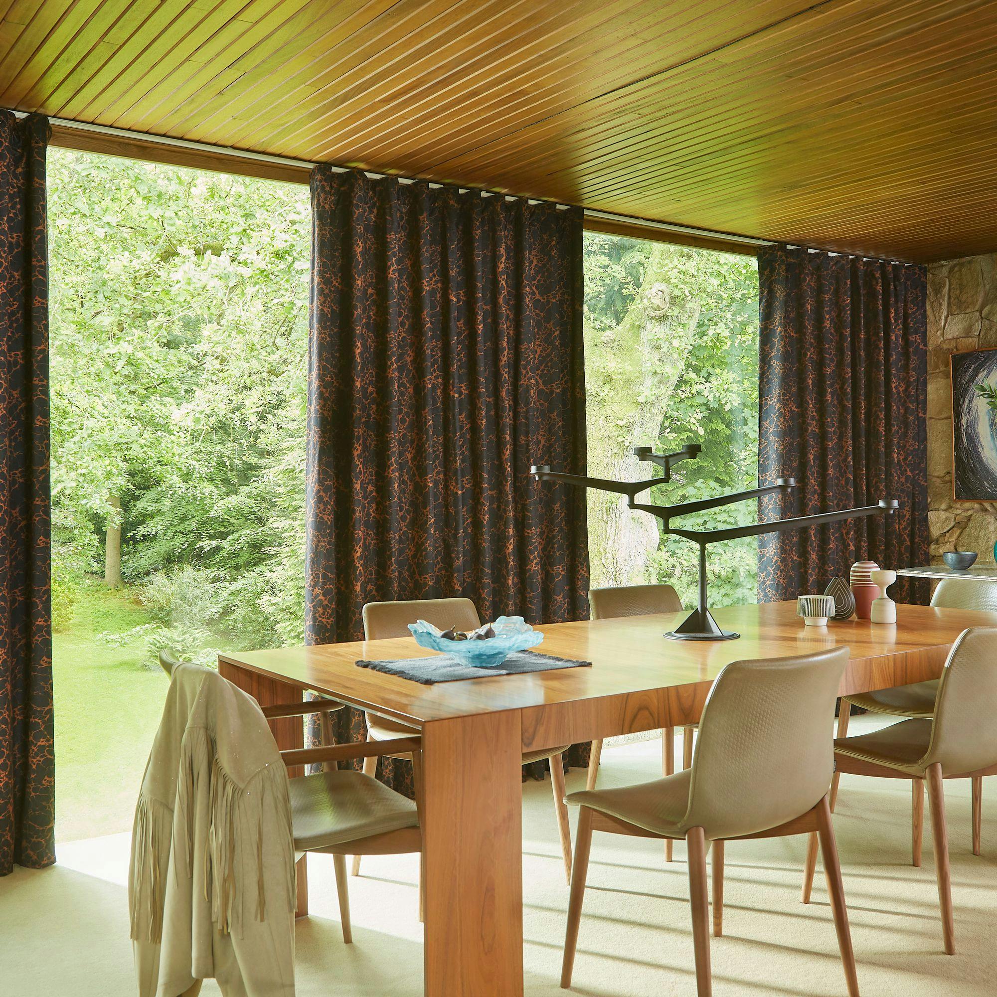 A wooden dining table with chairs surrounds decoratively arranged objects, including a sculptural lamp, near floor-to-ceiling windows with patterned curtains, overlooking a lush green garden.