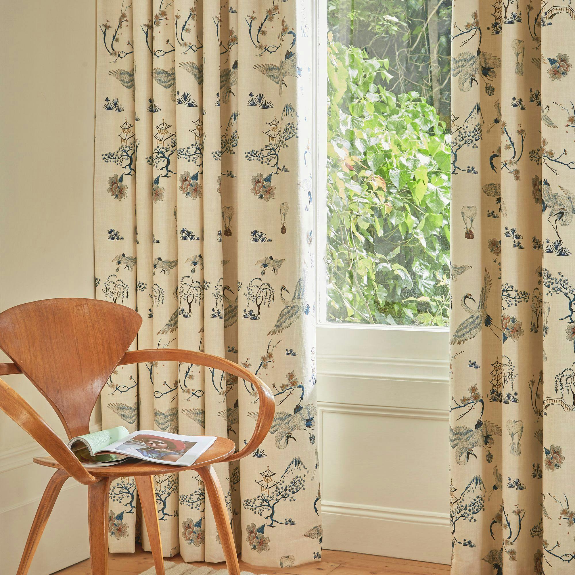 A wooden chair holding an open magazine sits in front of floral-patterned curtains in a room. Sunlight filters through a window with leafy greenery outside.