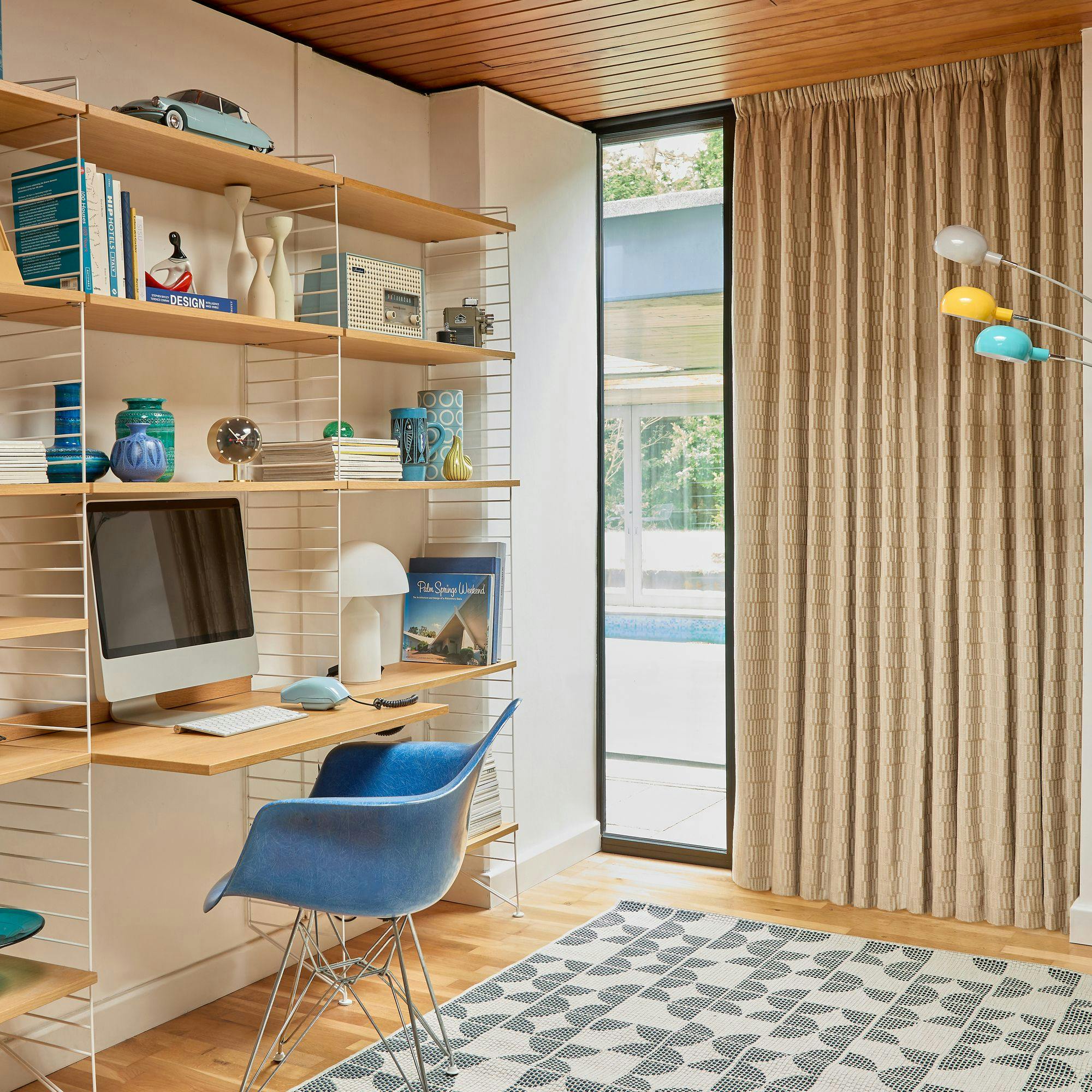 A home office setup with a computer on a wooden desk, surrounded by shelves holding books, decorative objects, and a toy car. A blue chair sits nearby, with floor-to-ceiling windows and curtains in the background.