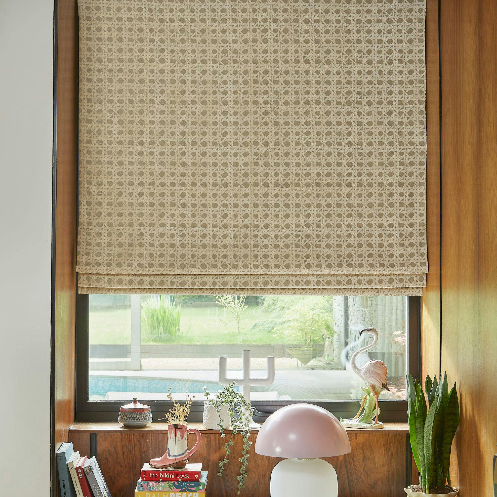 A patterned beige blind covers a window overlooking a garden. A wooden ledge holds books, plants, a pink lamp, and a small flamingo statue. Outside, greenery and a pool are visible.
