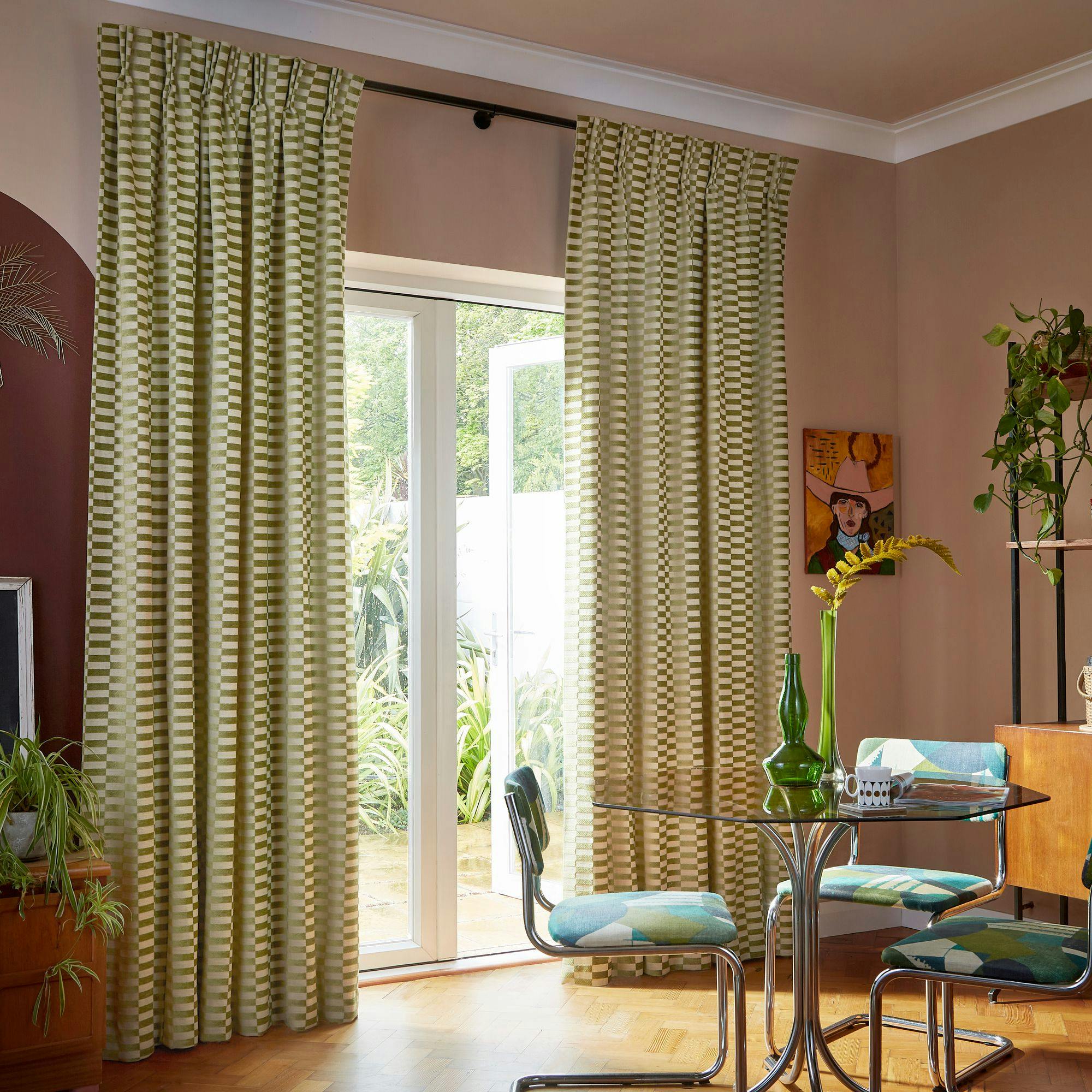 Curtains hang around a glass door, partially open to a garden view. A glass table with a vase and chairs is in the room, adorned with plants and a colorful painting.
