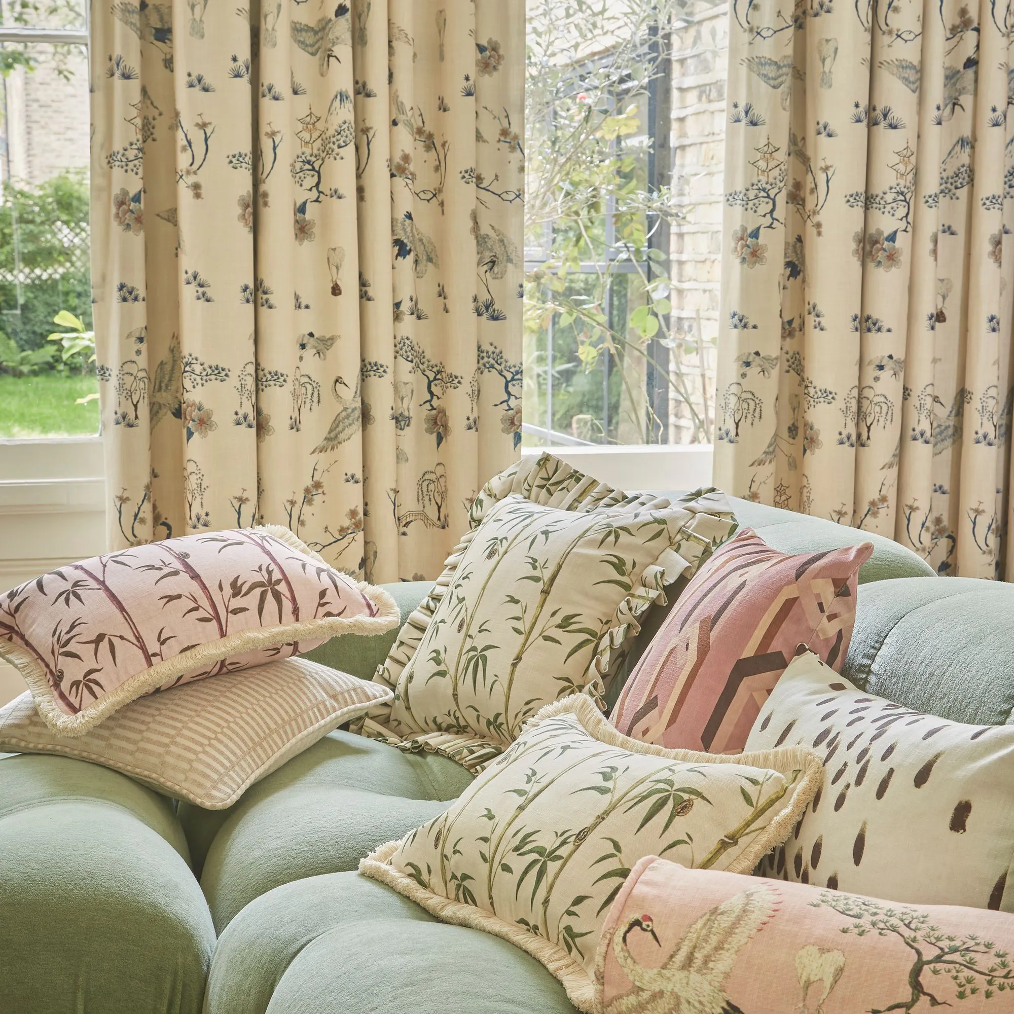 Cushions with botanical and animal prints rest on a tufted, light green sofa. Beige curtains with floral and bird patterns hang nearby, and sunlight streams through a window overlooking greenery.