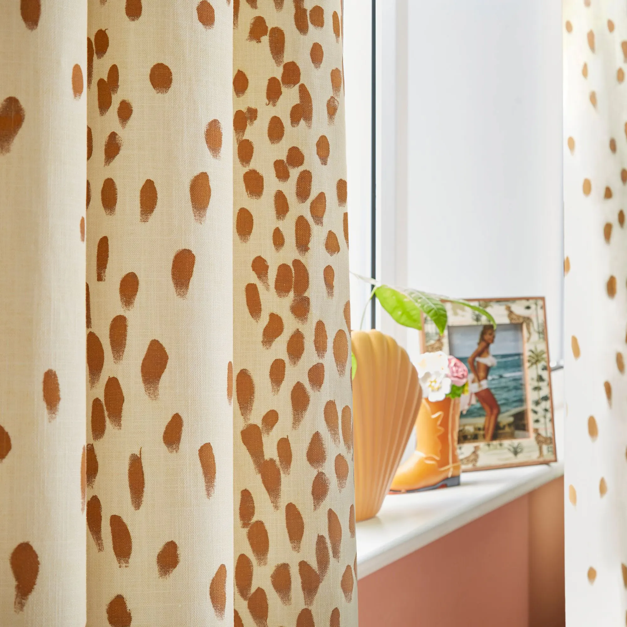 Curtains with brown polka dots hang beside a window. On the sill, a vase holds a plant and a framed beach photograph decorates the space.