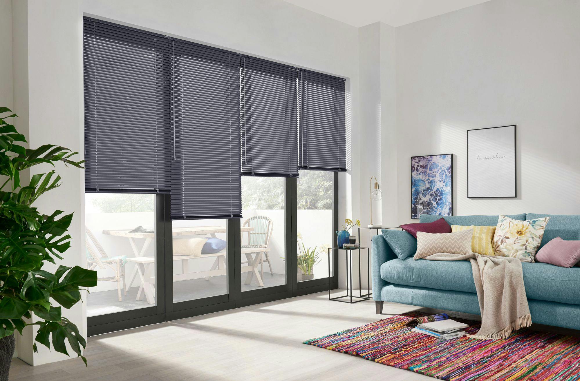 Blue sofa adorned with colorful pillows and a beige throw sits in a modern living room. Vertical blinds cover large glass doors. Wall art includes text: "breathe." A vibrant rug beneath.