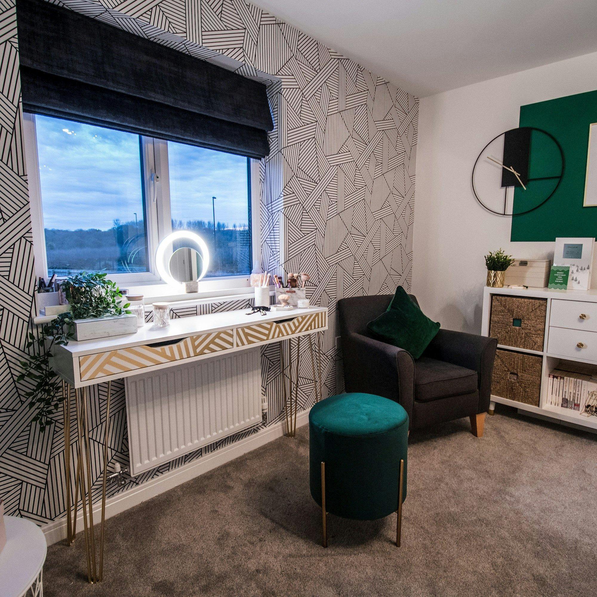 A stylish room features a vanity with a light-up mirror, cosmetics, and a plant, near a window. Adjacent is a cushy chair, ottoman, and modern wall decor in a geometrically patterned space.