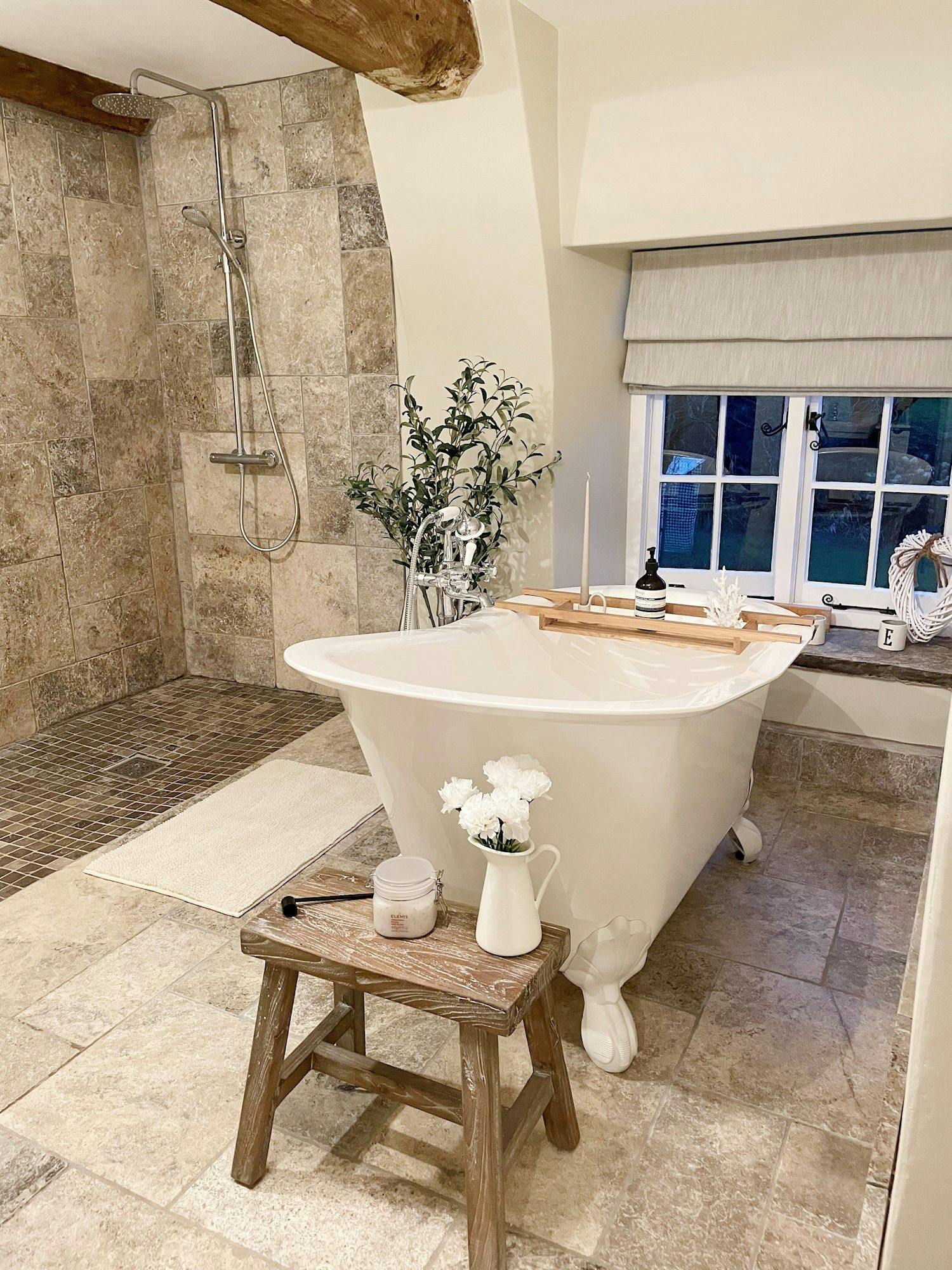 A white clawfoot bathtub sits beside a wooden stool holding a pitcher of flowers and a jar. Surrounding it, tiled walls and floor, a rain shower, and a window with a shade.