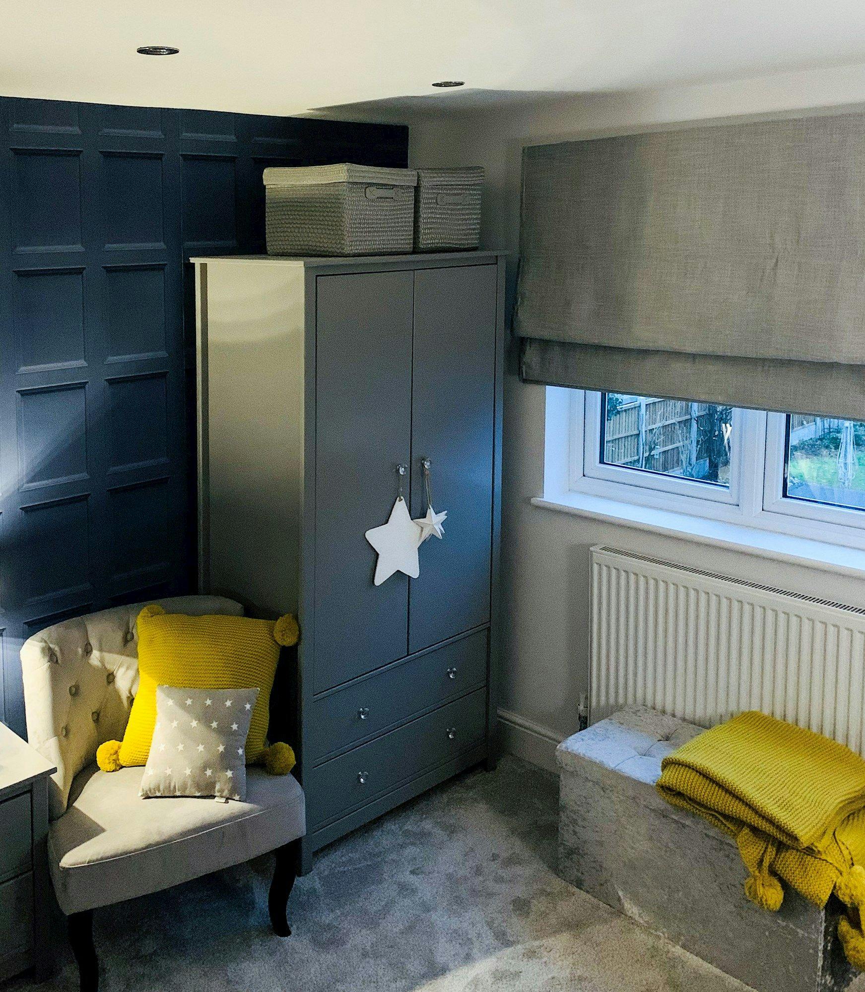A gray wardrobe with star decorations stands beside a tufted chair with yellow cushions in a room with a blue paneled accent wall and a window with closed gray blinds.