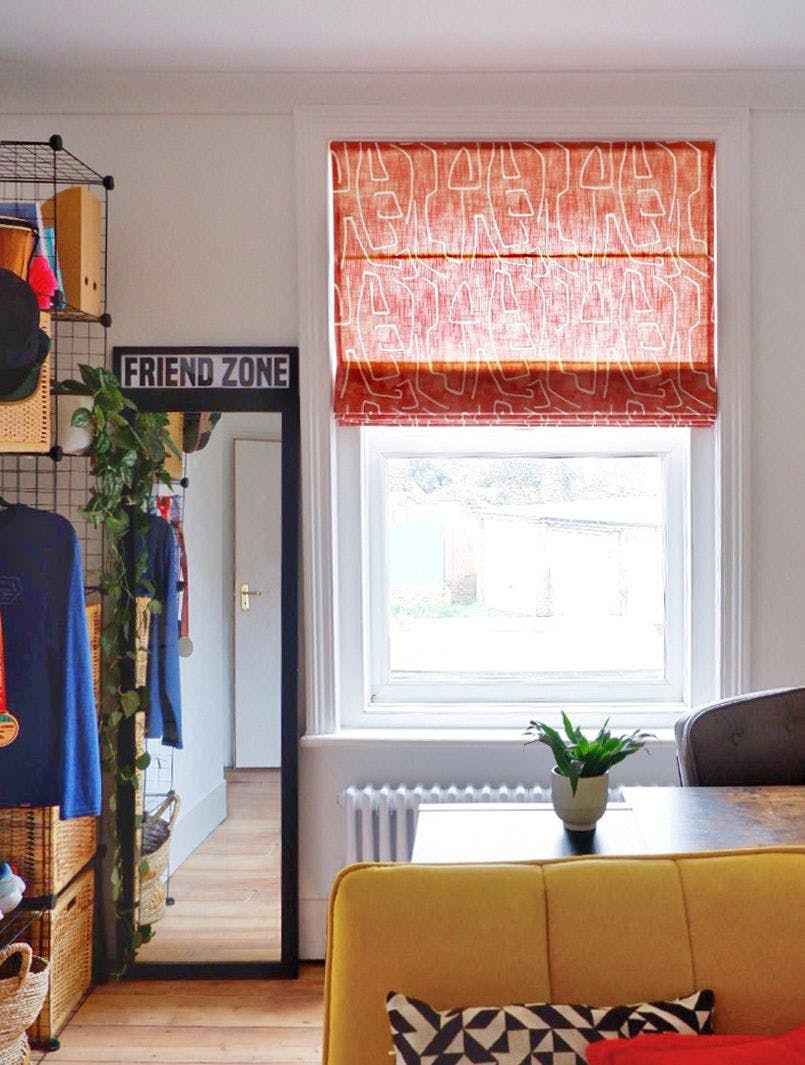 A tall mirror with a "FRIEND ZONE" sign leans against a wall; beside it, a window with orange blinds illuminates a yellow sofa, patterned pillow, and plant on a table.
