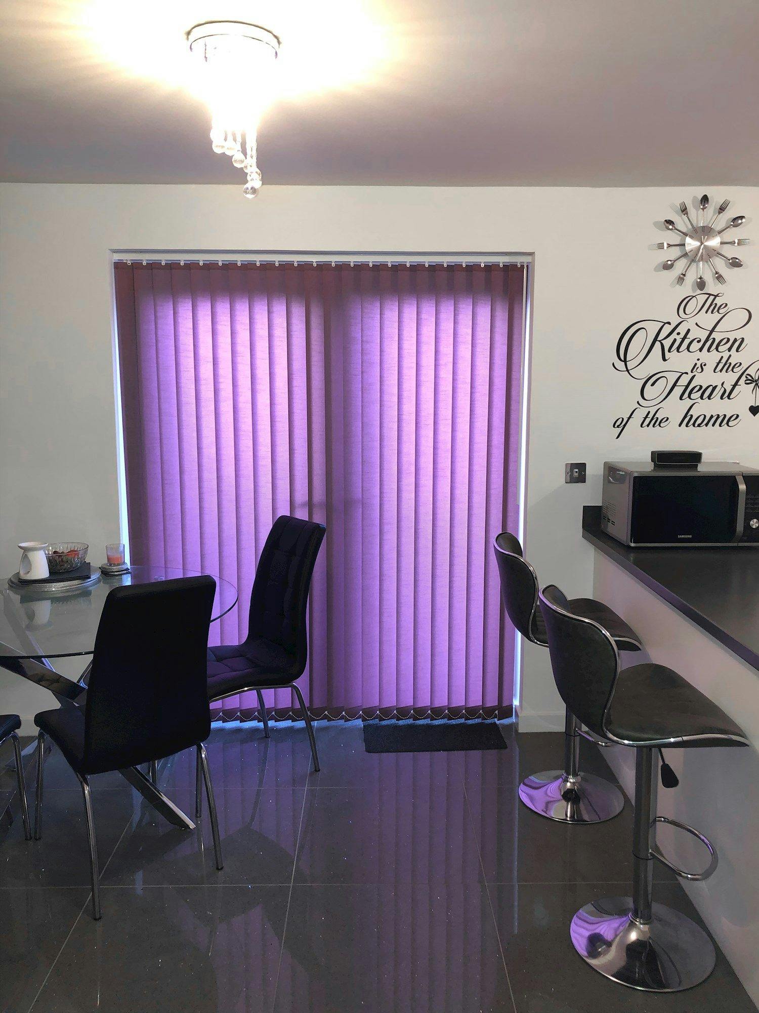 Dining table with black chairs sits on glossy tiled floor, next to purple blinds. A counter with bar stools is nearby. Wall text reads, "The Kitchen is the Heart of the home."