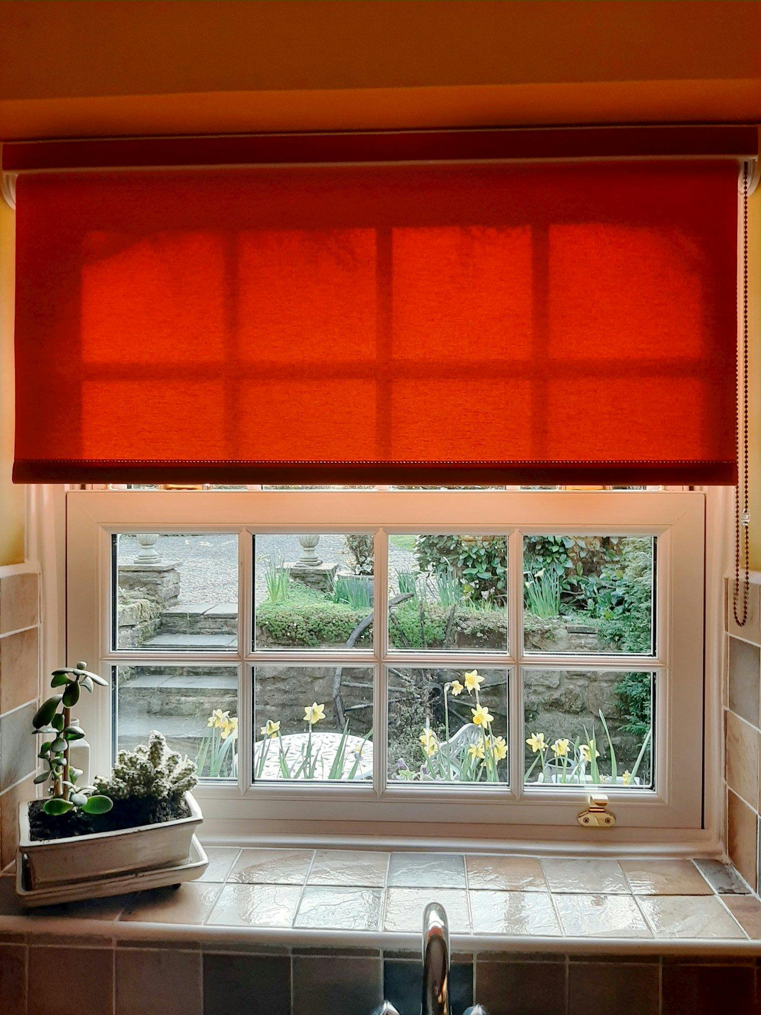 A kitchen window features a red roller blind partially rolled up, revealing a garden with daffodils and stone steps. A potted plant sits on the tiled windowsill.