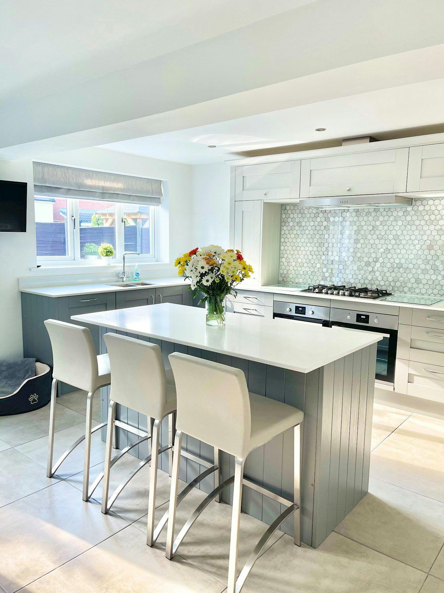 A kitchen island with a vase of flowers sits in a modern kitchen with white cabinets, light gray backsplash, bar stools, and a pet bed near the window.