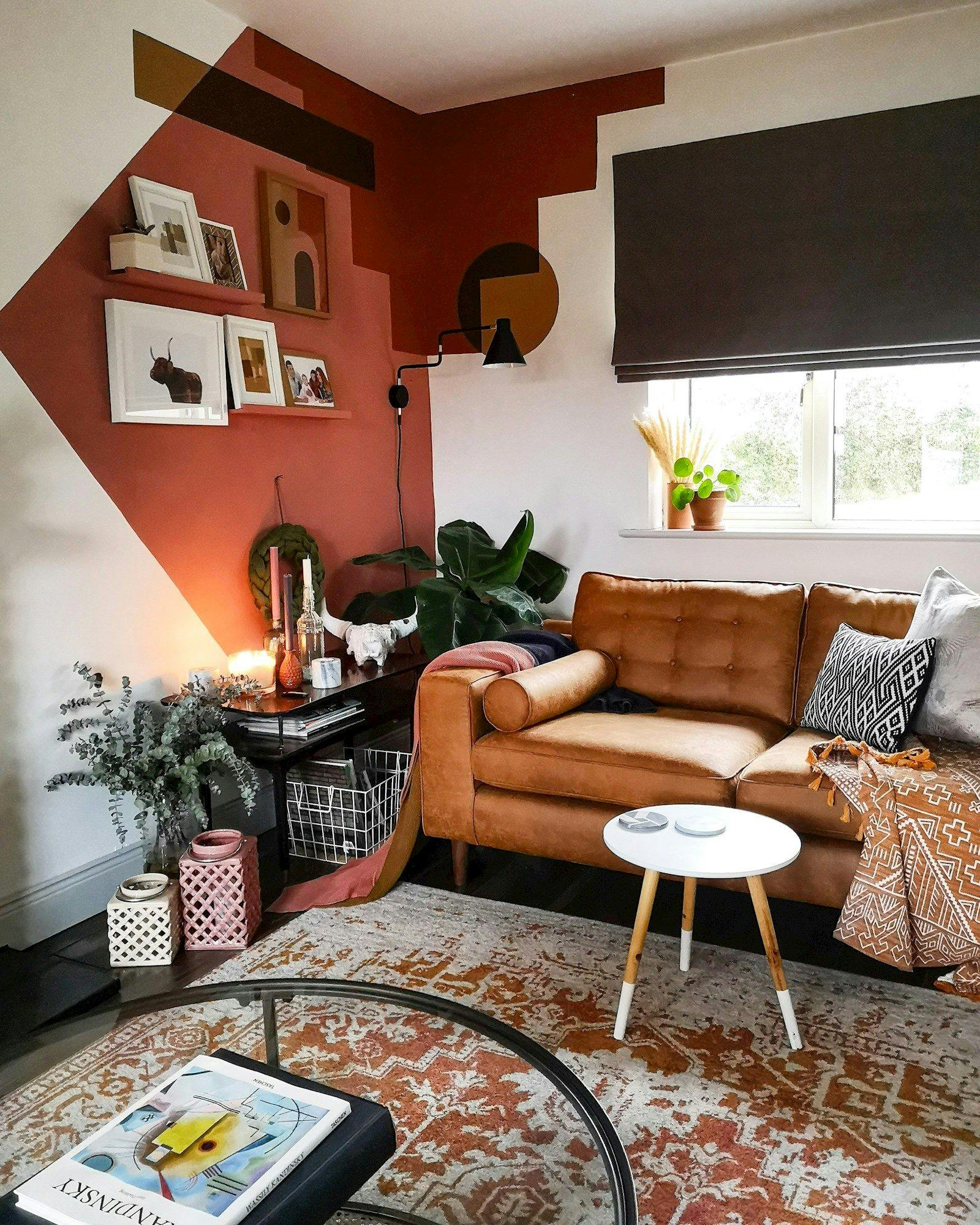A brown leather sofa with patterned cushions sits in a stylized living room. Geometric wall art decorates the red and white walls, and a window with plants provides natural light.