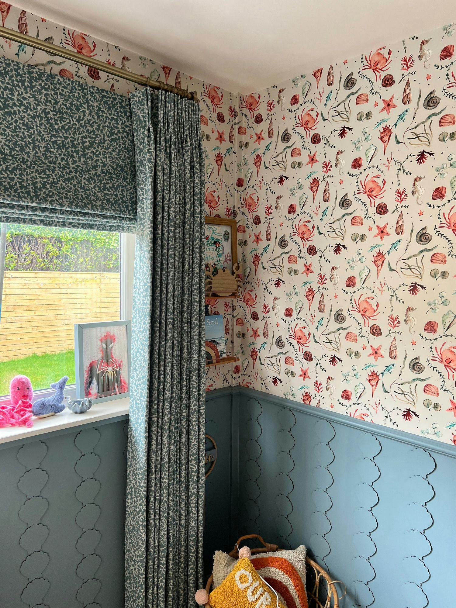 Curtains drape beside a window with blue floral patterns, set in a room with a lively sea-themed wallpaper of shells and coral. Soft toys sit on the windowsill, enhancing coziness.