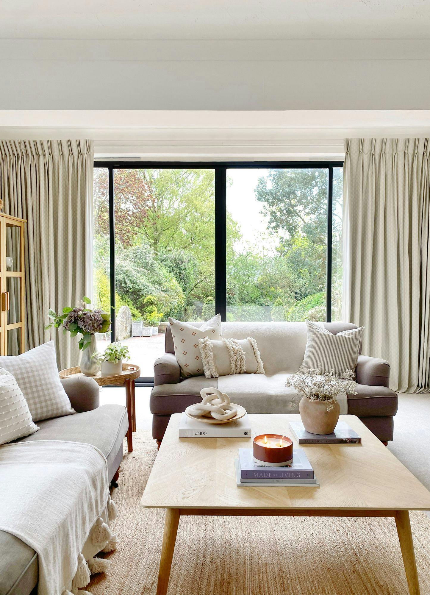 A cozy living room features two sofas with decorative pillows, a wooden coffee table with books and a lit candle, and large windows overlooking a lush garden. The books are titled "at 100" and "MADE FOR LIVING".
