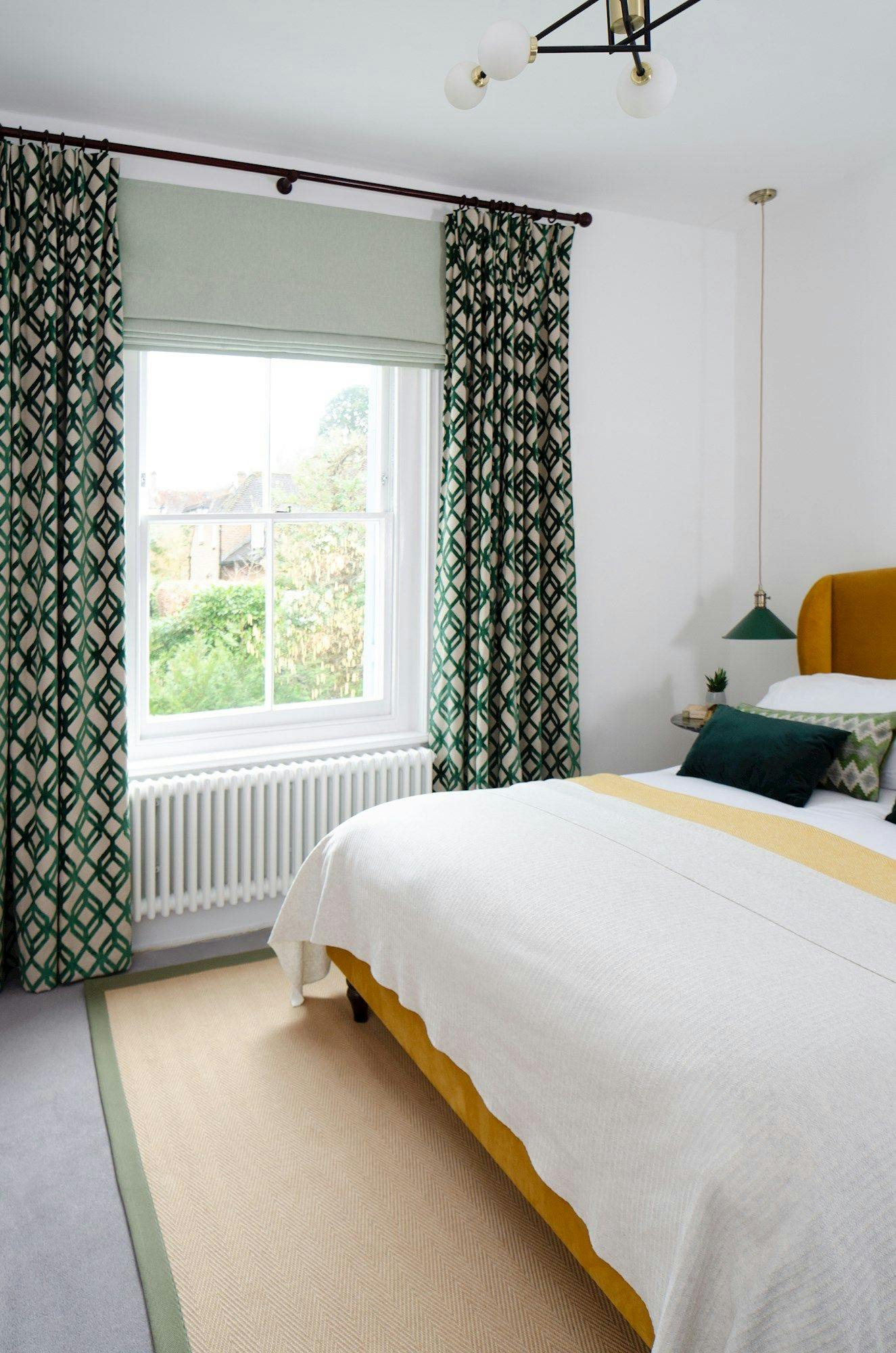 A bed with white and mustard bedding stands beside a large window with patterned green curtains, in a bright bedroom with a grey carpet and pendant light.