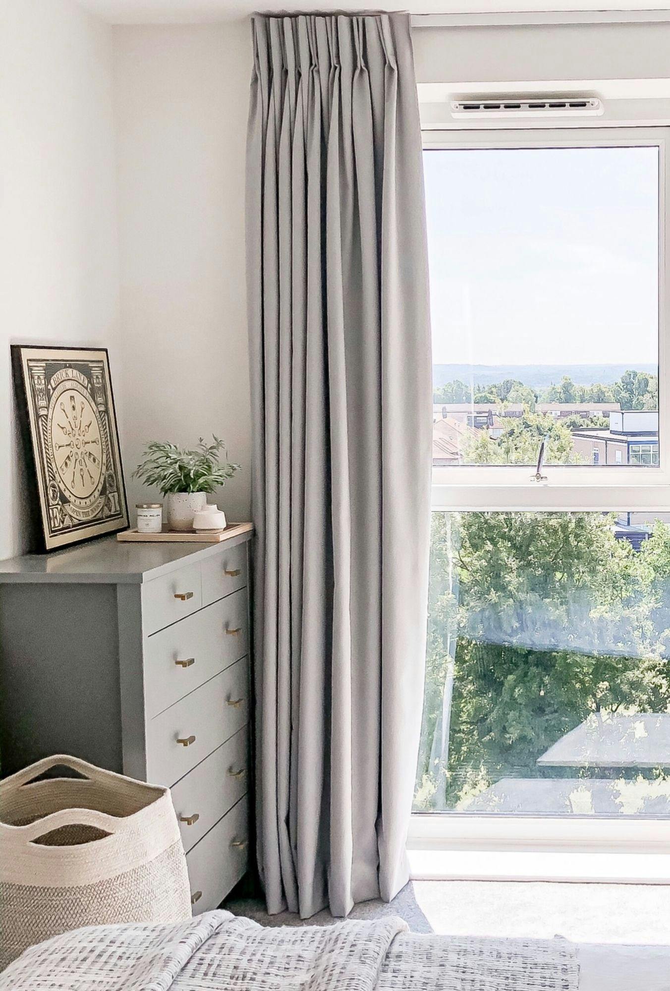 A dresser holds a decorative plant and framed artwork beside a window with closed curtains. A woven basket is nearby within a bright, airy room overlooking greenery outside.
