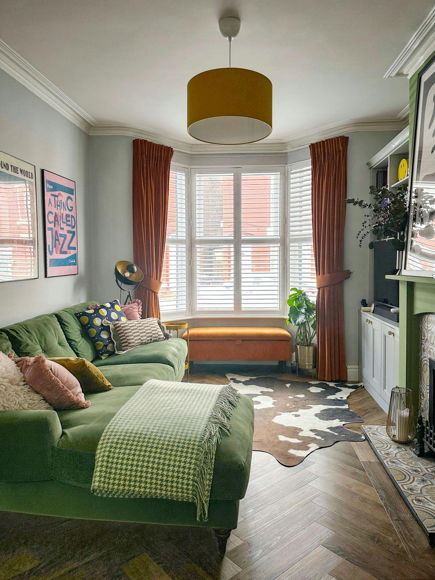 Green sofa with cushions and blanket facing a large window adorned with orange curtains. A cowhide rug lies on the wooden floor. Wall art reads "A Thing Called Jazz."