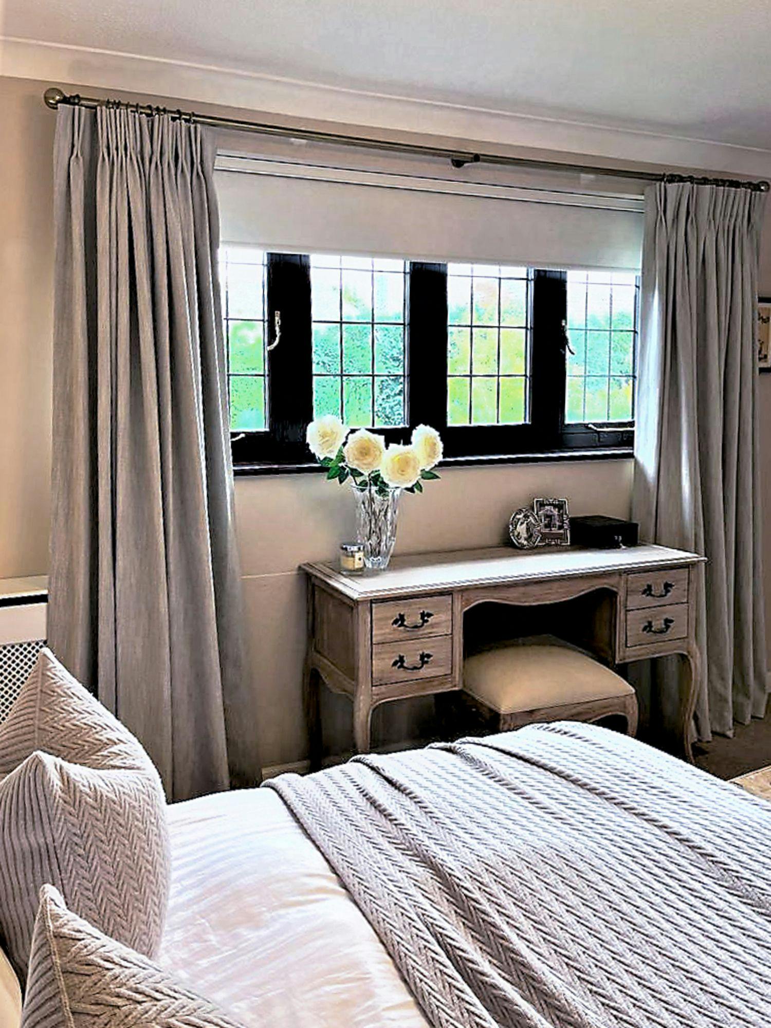 A wooden dressing table with drawers holds a vase of white flowers and a small framed picture against a window with closed gray curtains, in a cozy bedroom setting.