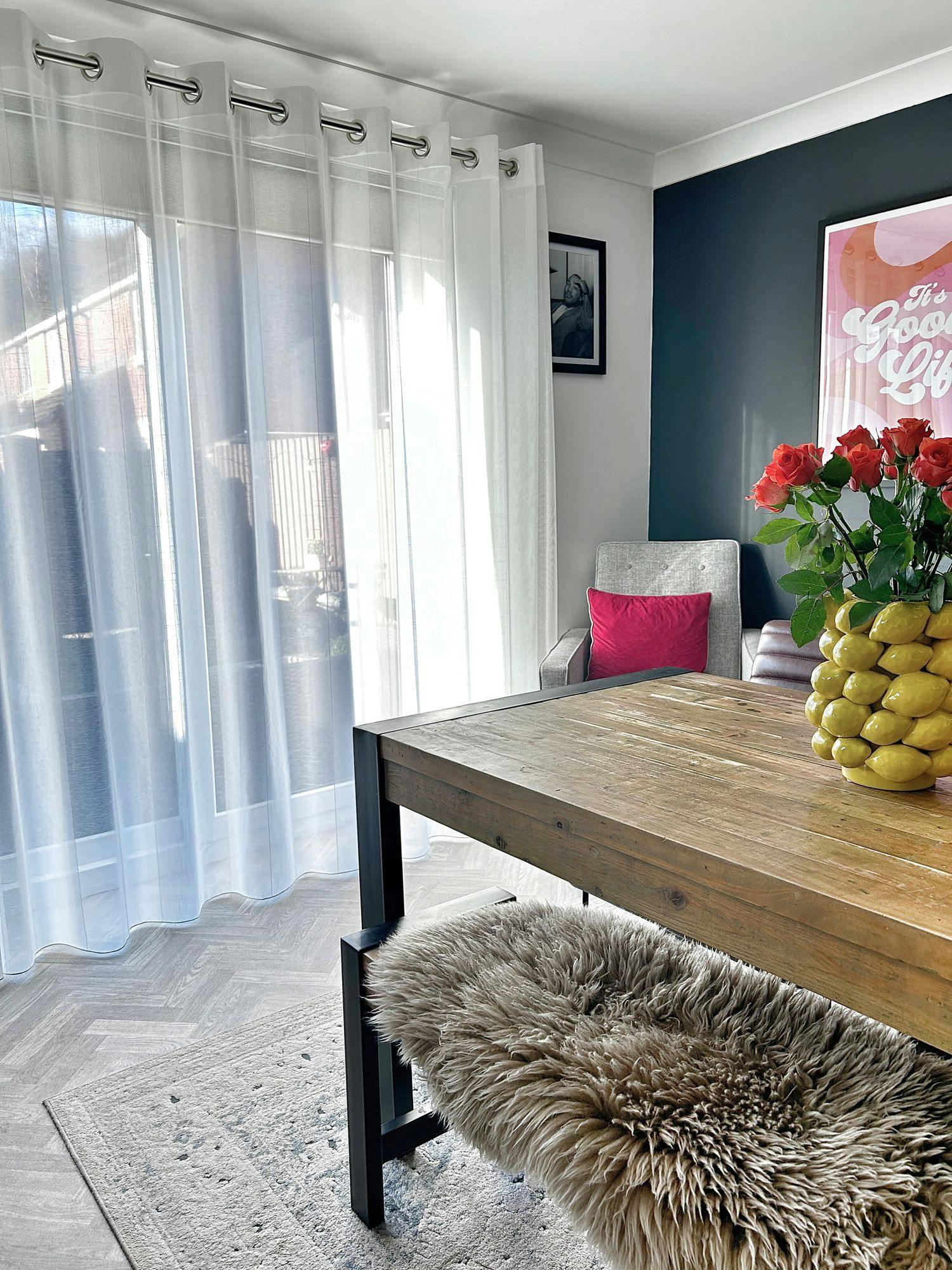 A wooden table with a vase of red roses sits on a light rug. A sheepskin-covered bench is beneath the table. "It's a Good Life" is written on a colorful wall poster near sheer curtains.