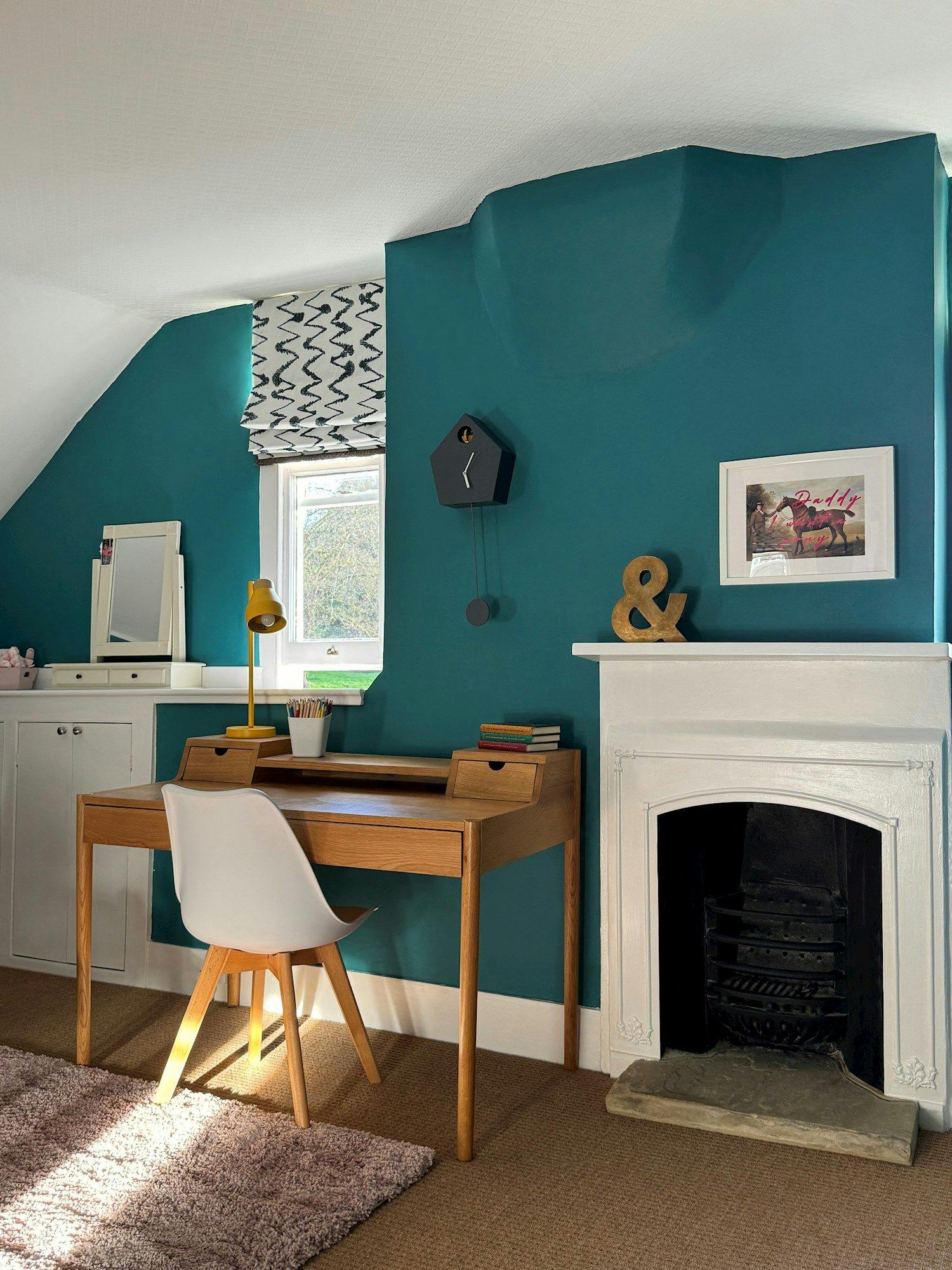 A wooden desk with a white chair sits on carpet next to a teal wall featuring a wall clock, fireplace, and framed artwork reading "Daddy & Daddy." A window illuminates the scene.