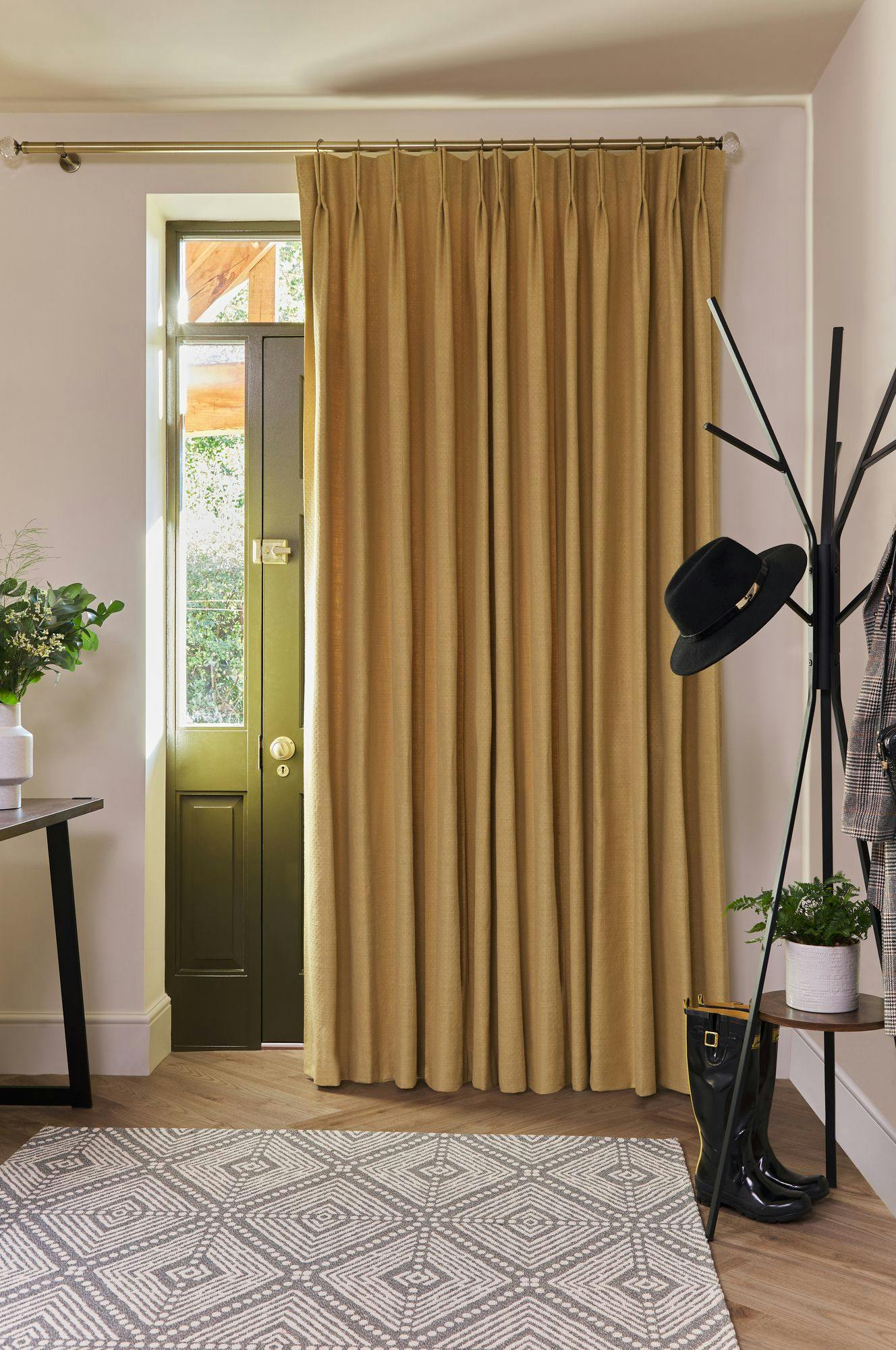 A beige curtain hangs across a door in a cozy room. Nearby, a black hat rests on a coat rack, and a table holds a potted plant. A patterned rug decorates the floor.