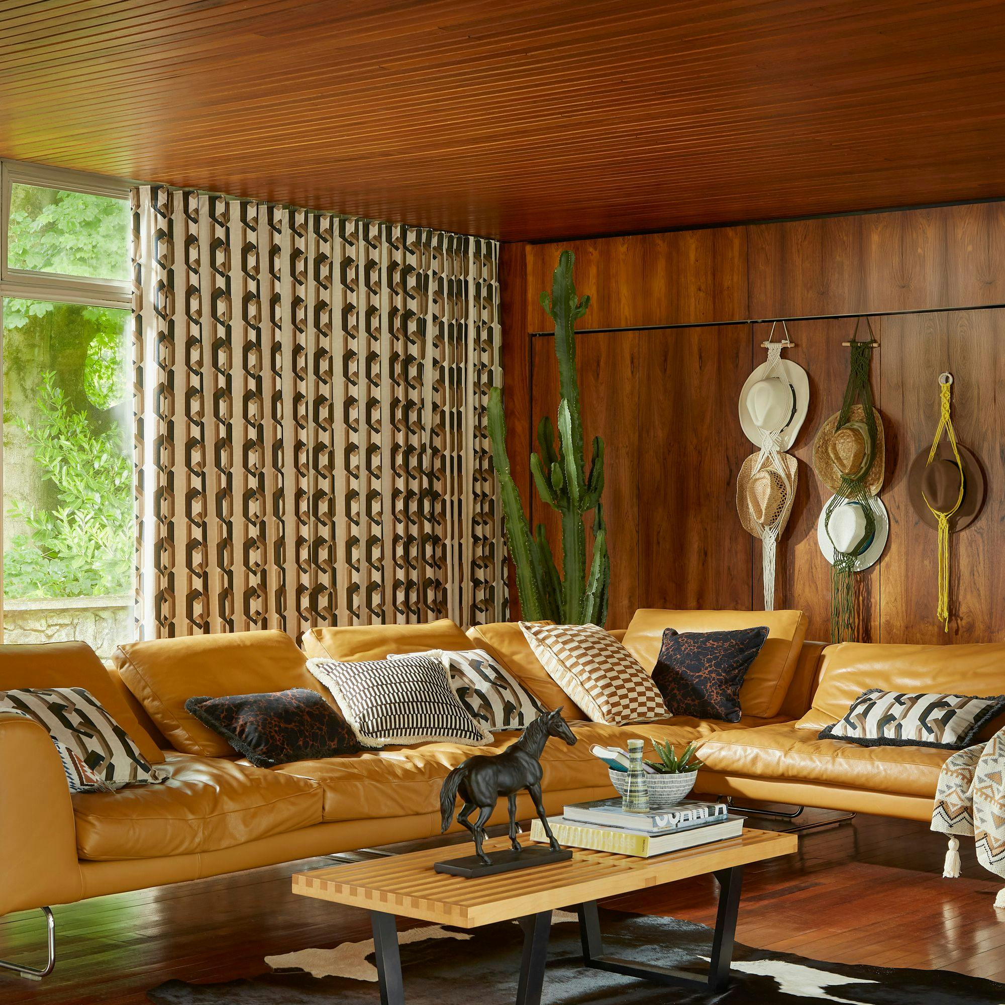 A cozy living room features a mustard-yellow leather sofa adorned with patterned pillows. A wooden wall with hanging hats and a cactus stand nearby. A table displays a horse figurine and books.