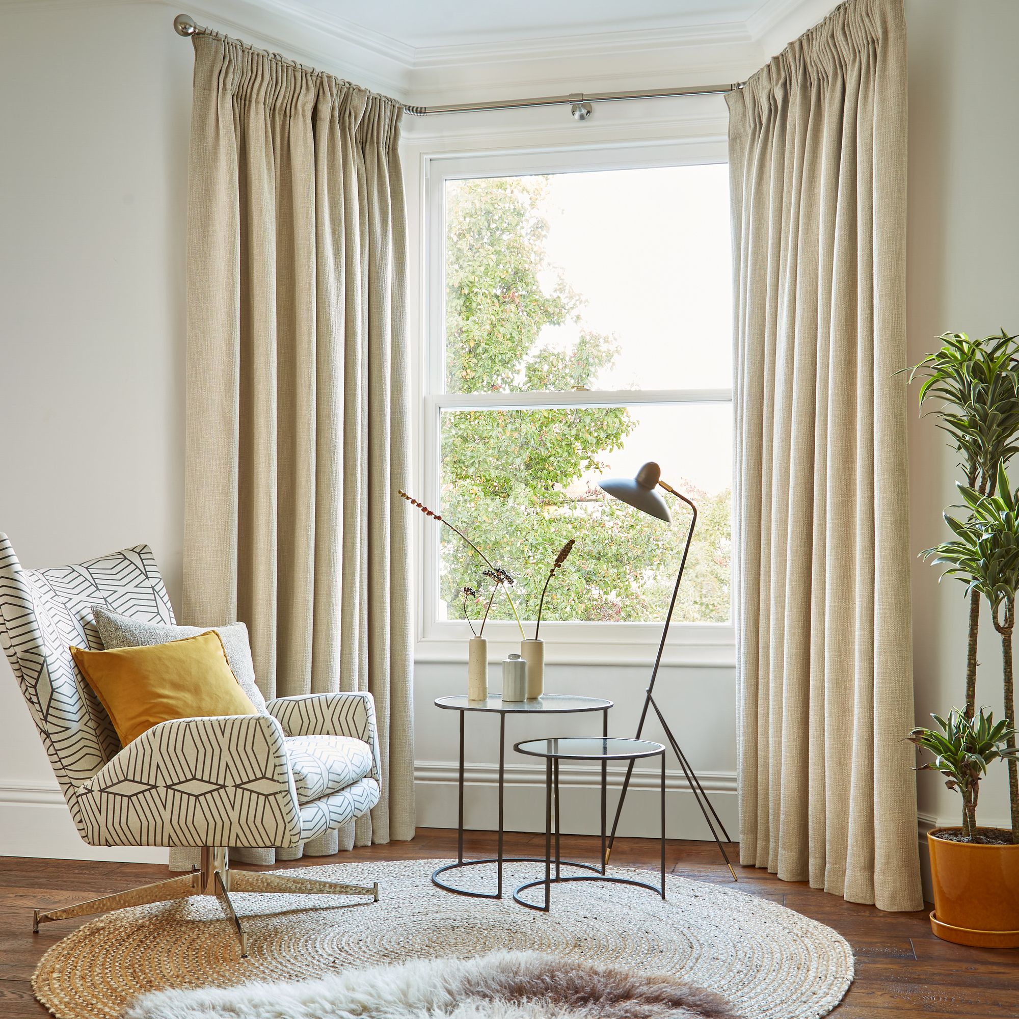 A patterned armchair sits beside a round, nested coffee table with vases, illuminated by a modern floor lamp. Beige curtains frame a window overlooking greenery, enhancing the cozy room.