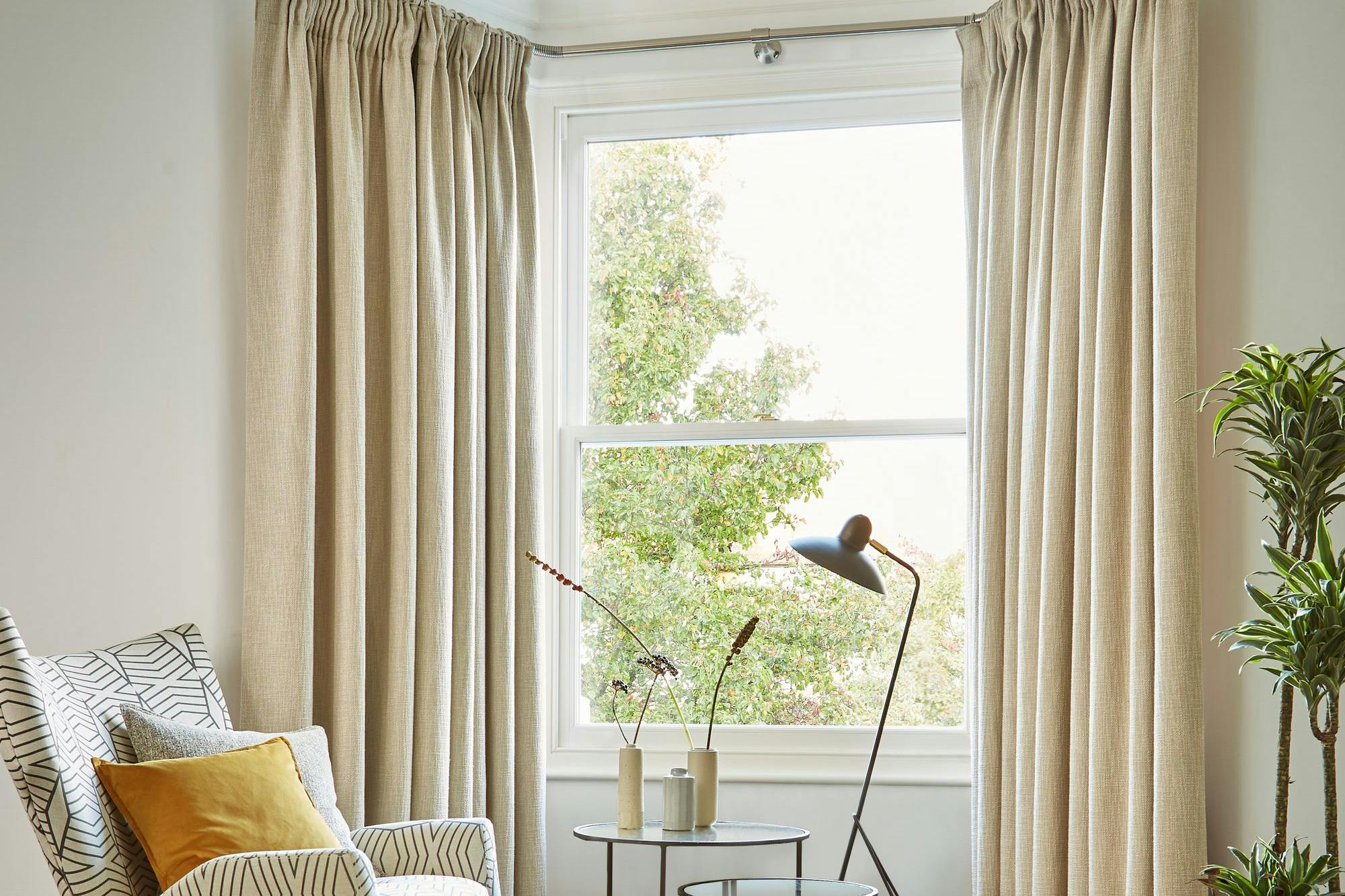 A patterned armchair sits beside a round, nested coffee table with vases, illuminated by a modern floor lamp. Beige curtains frame a window overlooking greenery, enhancing the cozy room.