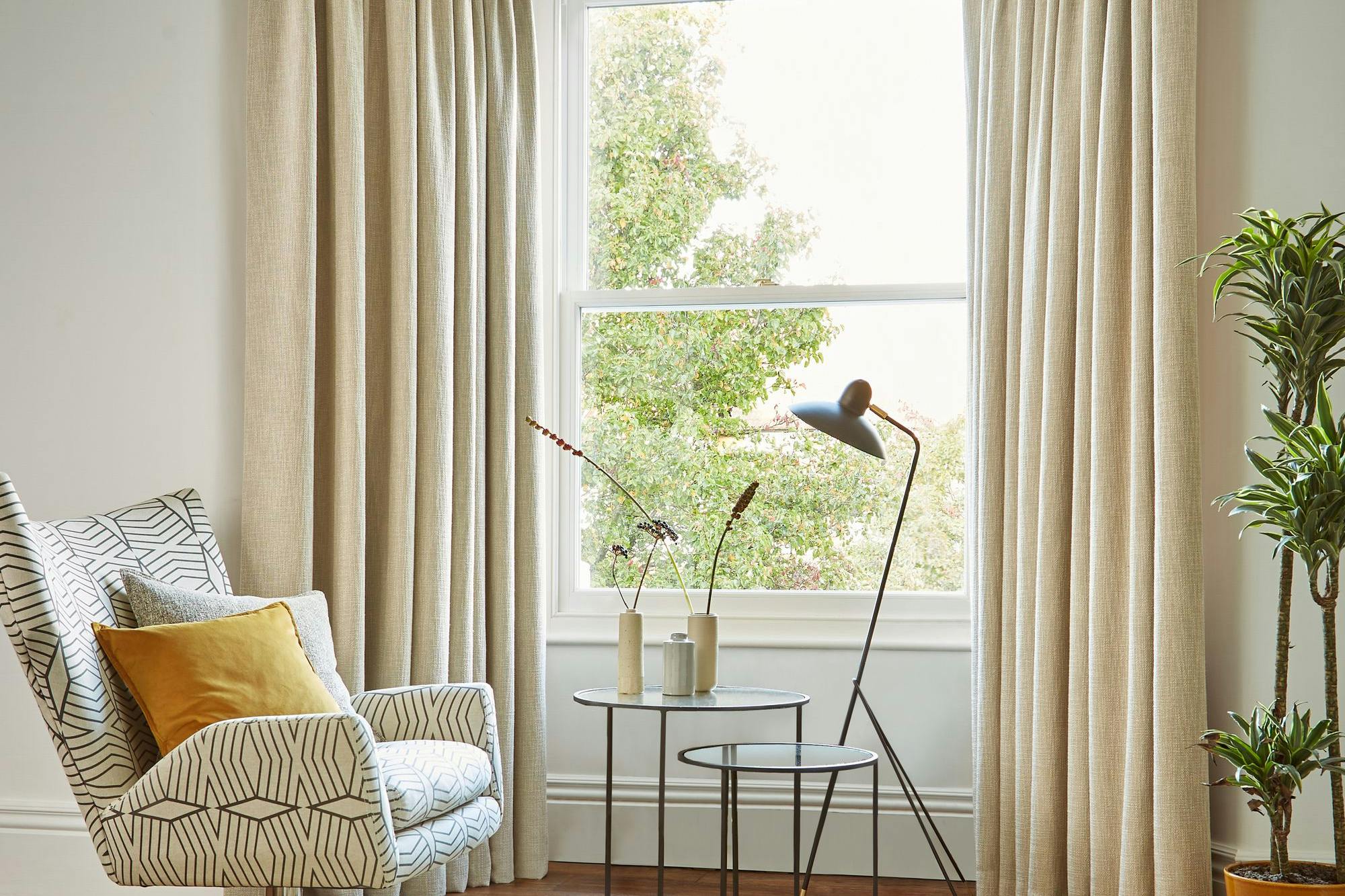 A patterned armchair sits beside a round, nested coffee table with vases, illuminated by a modern floor lamp. Beige curtains frame a window overlooking greenery, enhancing the cozy room.