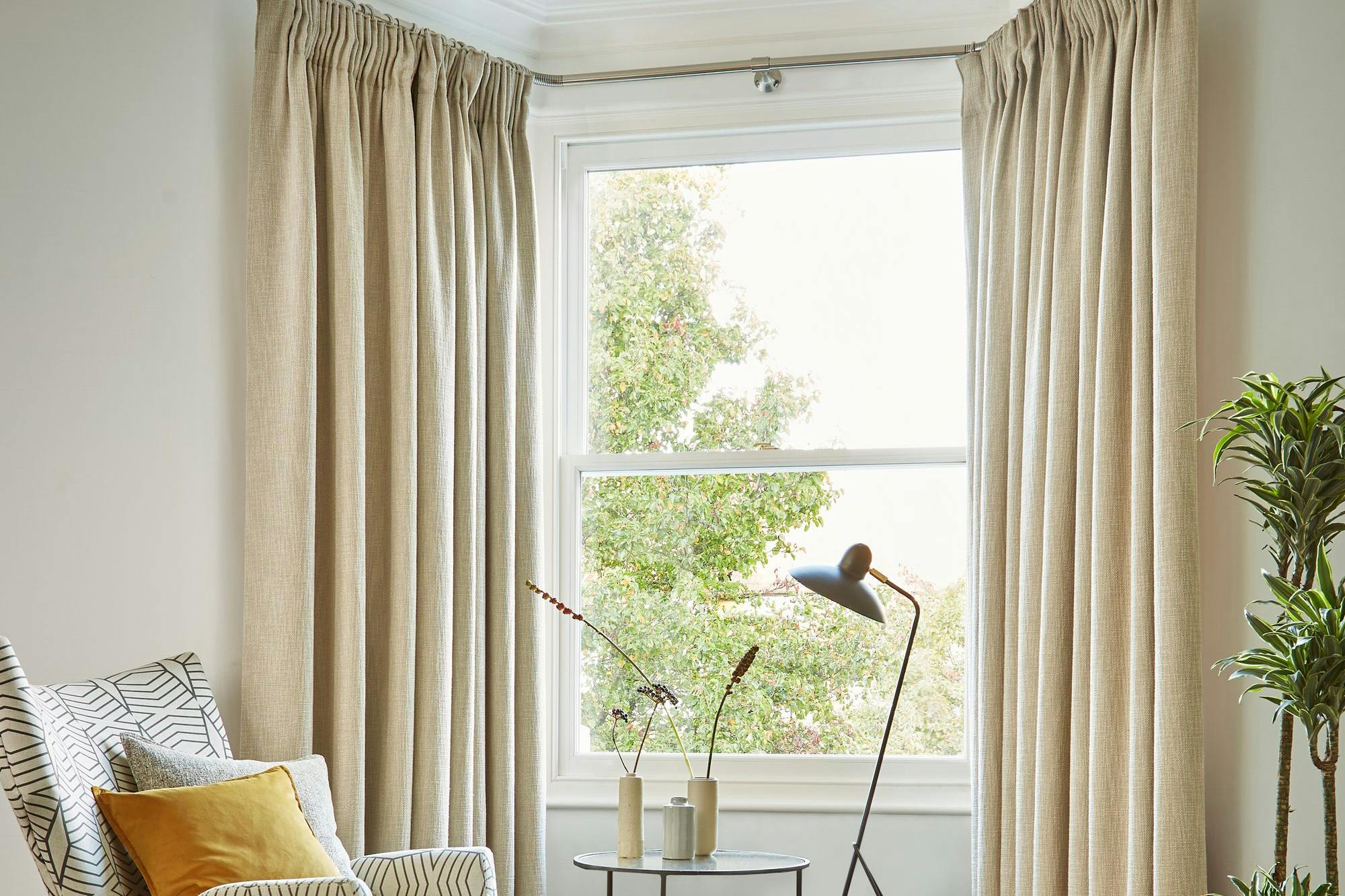 A patterned armchair sits beside a round, nested coffee table with vases, illuminated by a modern floor lamp. Beige curtains frame a window overlooking greenery, enhancing the cozy room.