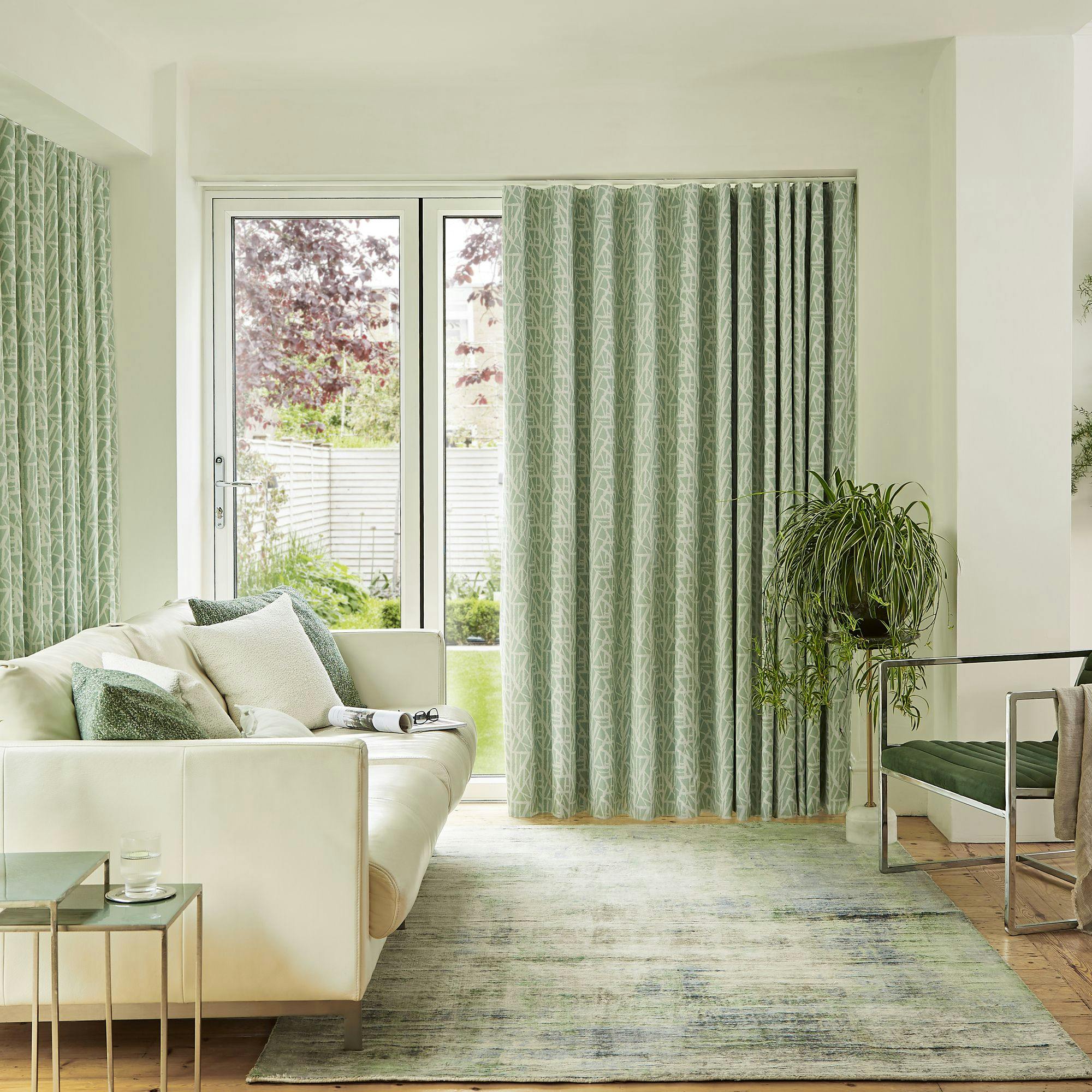 A white sofa with green pillows rests beside large, glass patio doors covered by patterned curtains; a plant and chair sit nearby in the bright, airy living room.