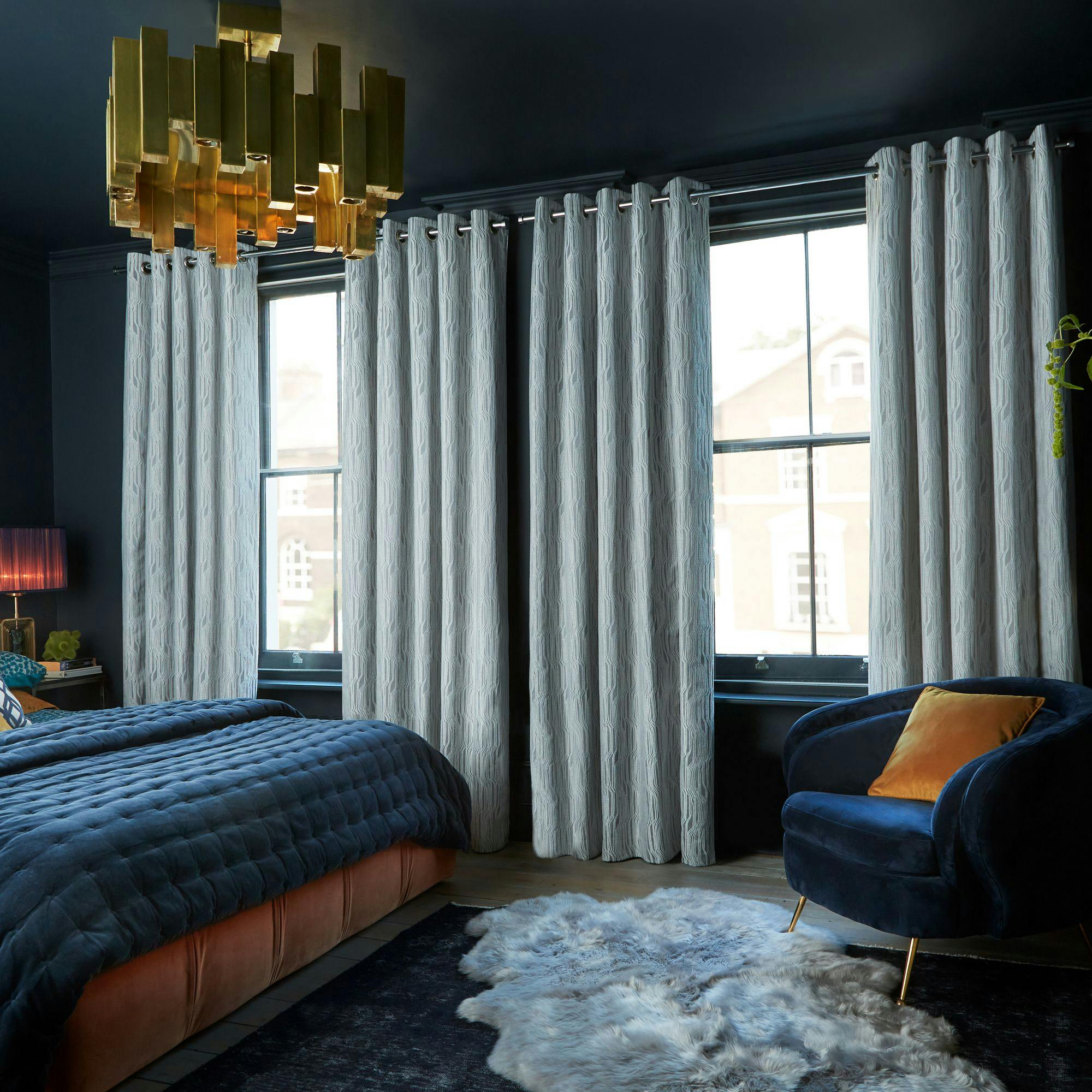 A bedroom with a dark blue bed and armchair, orange cushion, white curtains framing two windows, fluffy gray rug, golden geometric ceiling light, and dimly lit lamp on a bedside table.