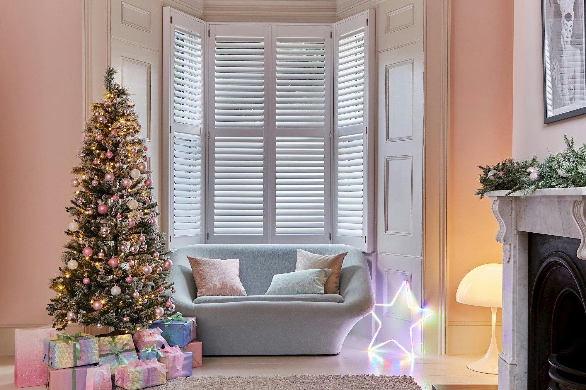 A decorated Christmas tree with pink ornaments stands beside a sofa. Presents are arranged underneath. The room features white shutters, a glowing star light, and a decorated mantle.