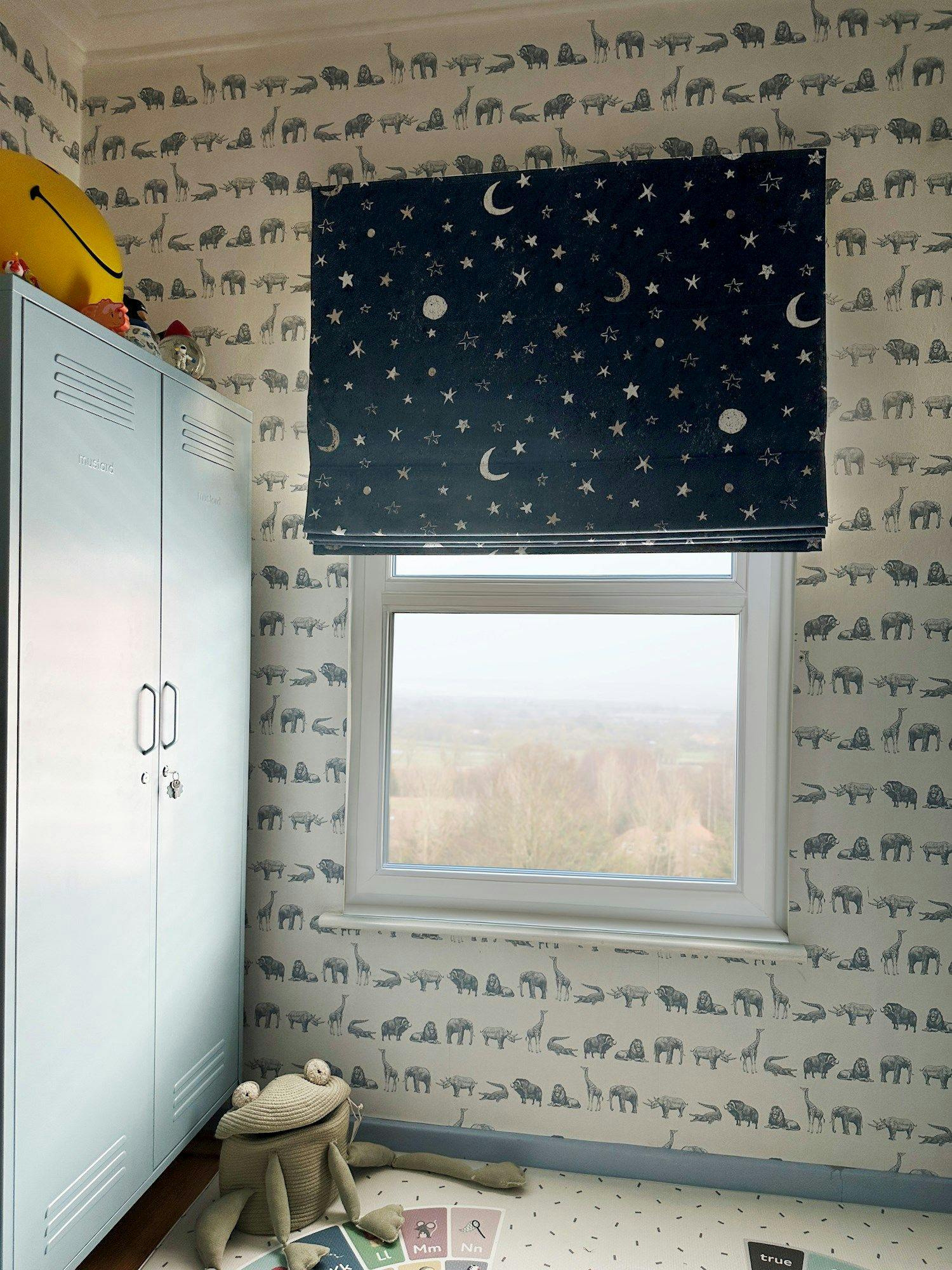 A room features a large window with starry night-patterned blinds, animal-printed wallpaper, a light blue locker labeled "Timbalo," and a plush frog toy on a speckled floor.