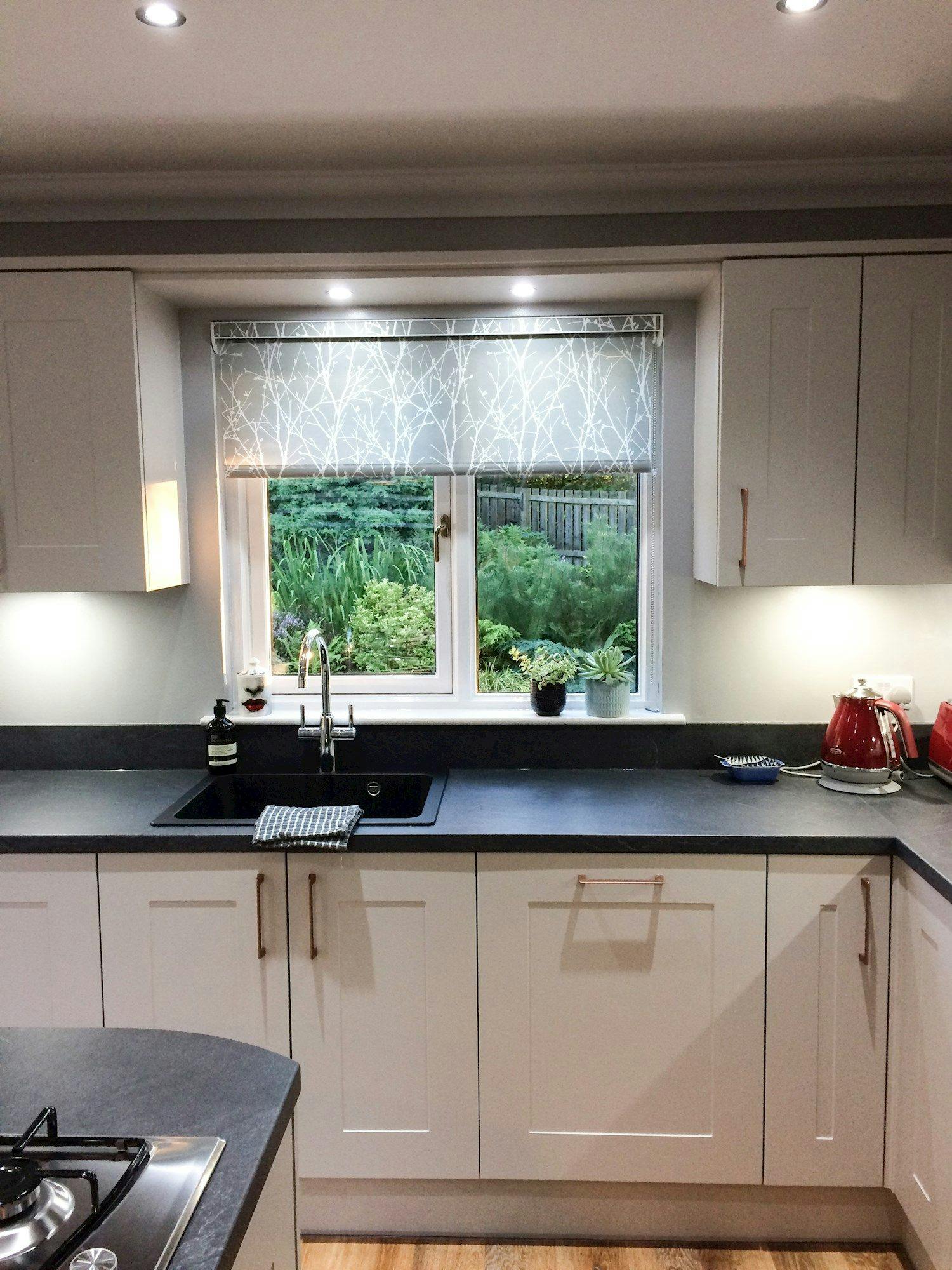 A modern kitchen features white cabinets, a black sink with a towel, and a window overlooking a garden. Appliances and plants adorn the countertop, under a patterned blind.