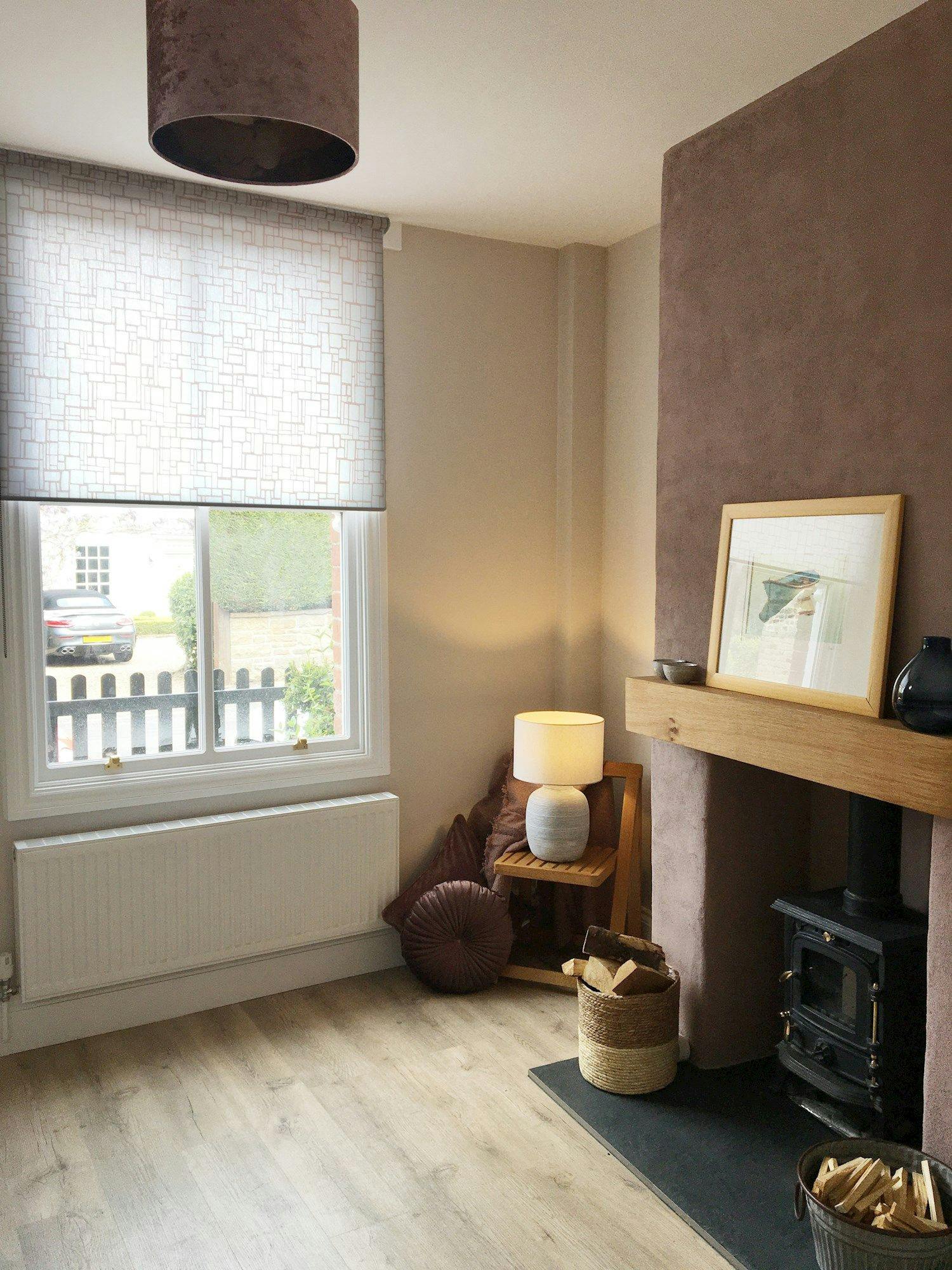 A cozy living room features a wood-burning stove with a basket of logs, a lamp on a small table, and a soft chair, all under soft natural light from a window.