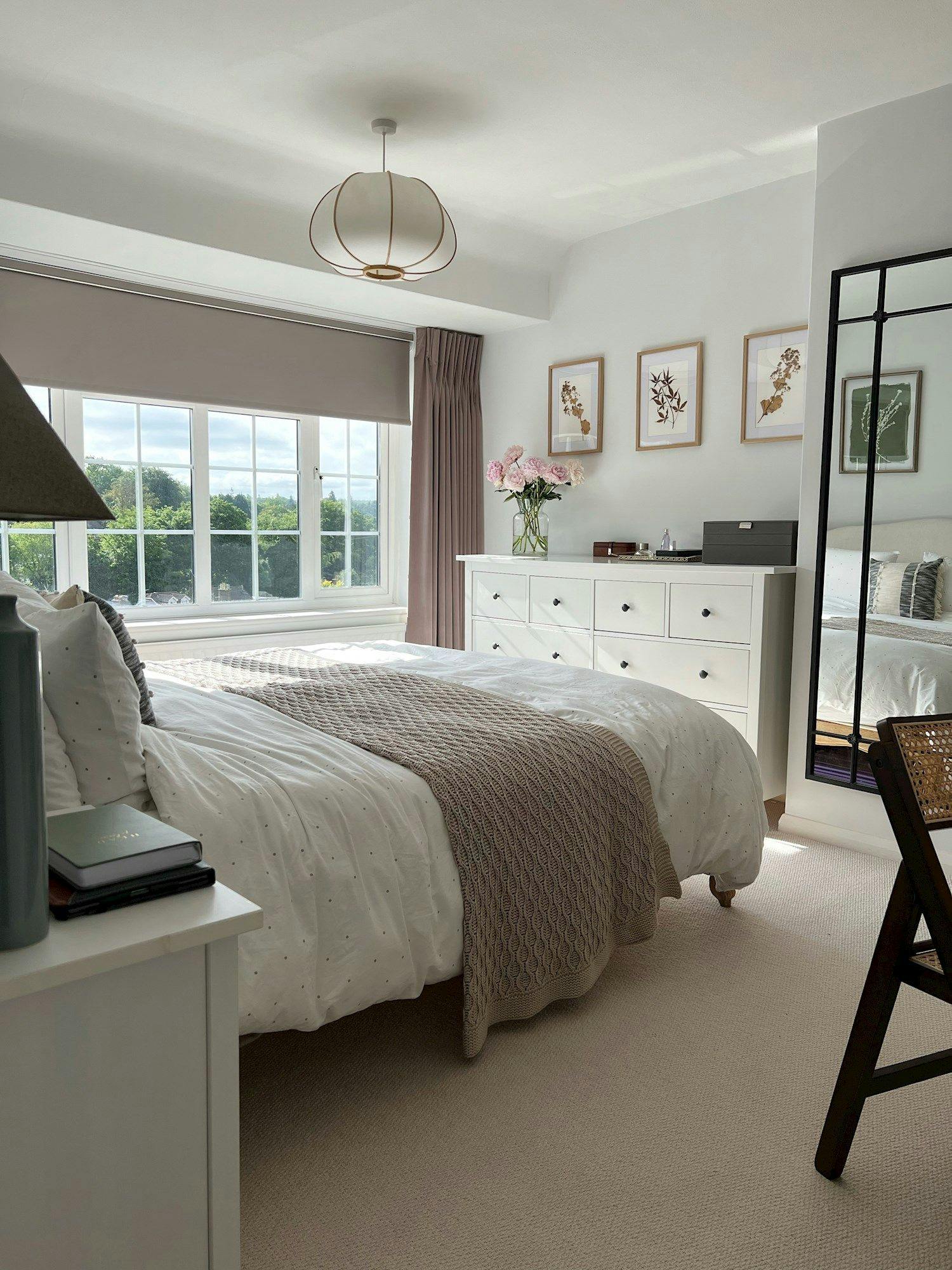 Bedroom with a neatly made bed, gray blanket, white duvet. Dresser with floral decor, window with green view. Three framed botanical prints adorn the white walls.