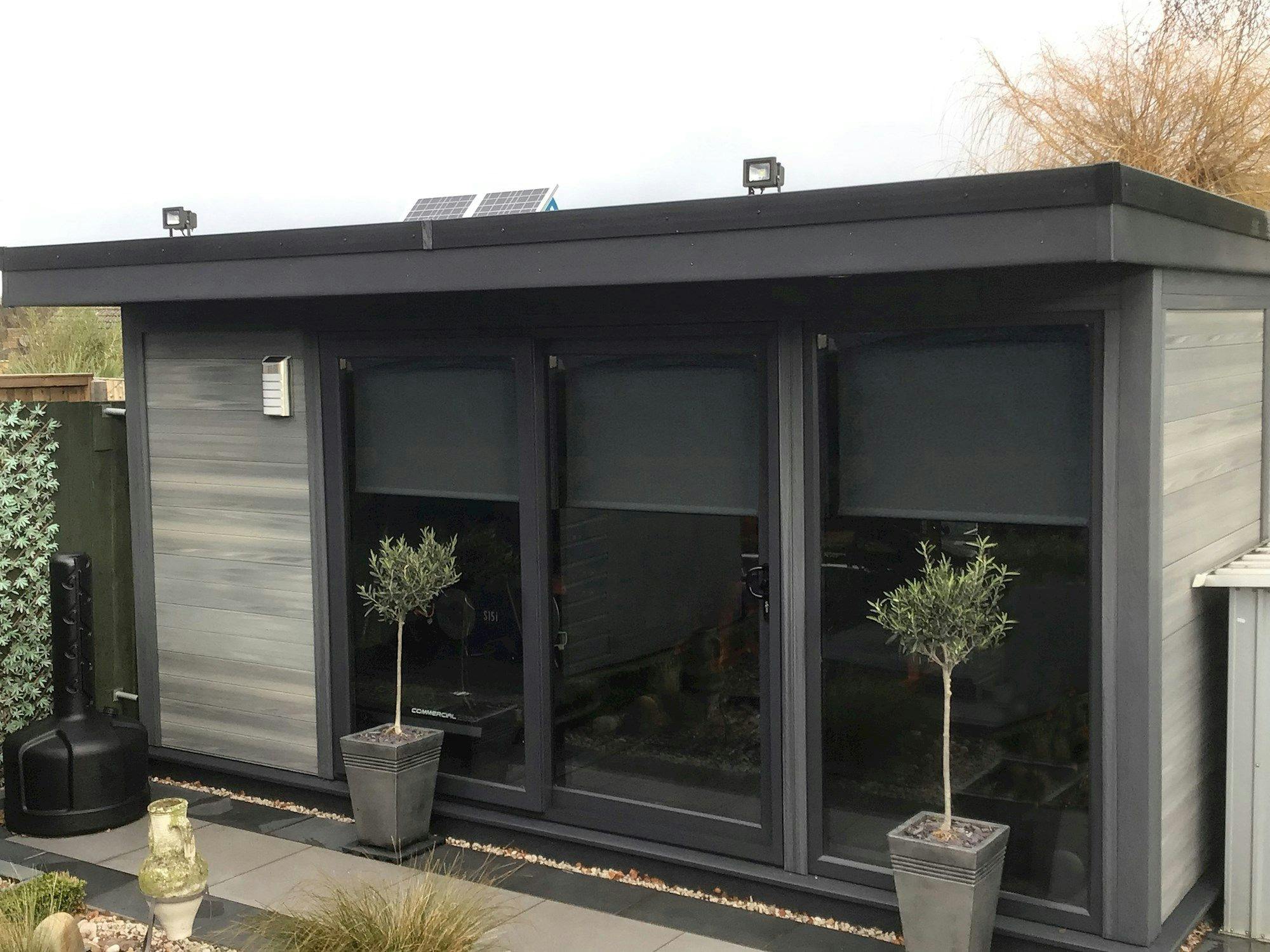 A modern garden shed with dark-framed sliding glass doors, flanked by small potted trees; solar panels and lights adorn its flat roof, set in a landscaped yard.